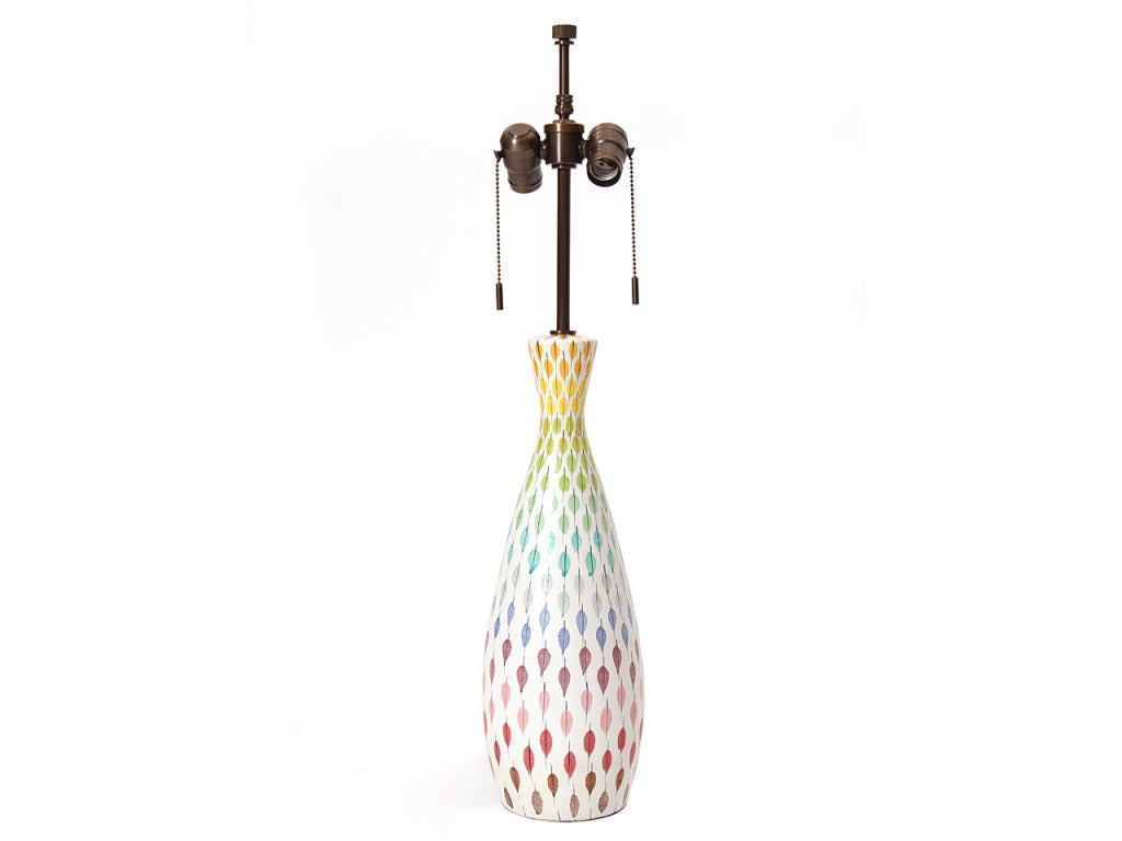 A hand thrown white ceramic table lamp with a repeating multi-color rainbow leaf motif.