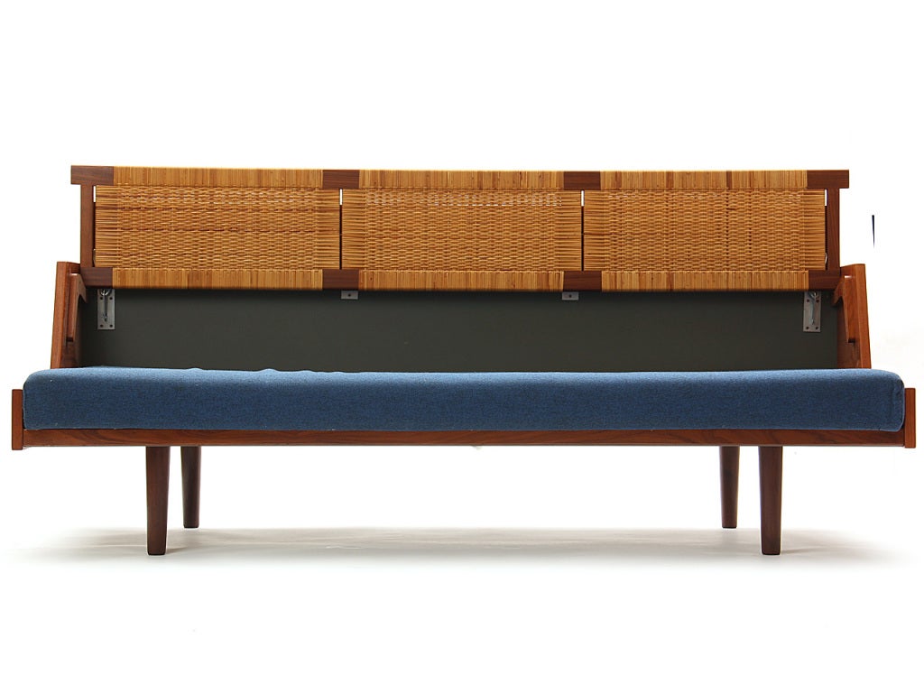 A convertible teak sofa daybed with a caned storage backrest, with an upholstered seat cushion.