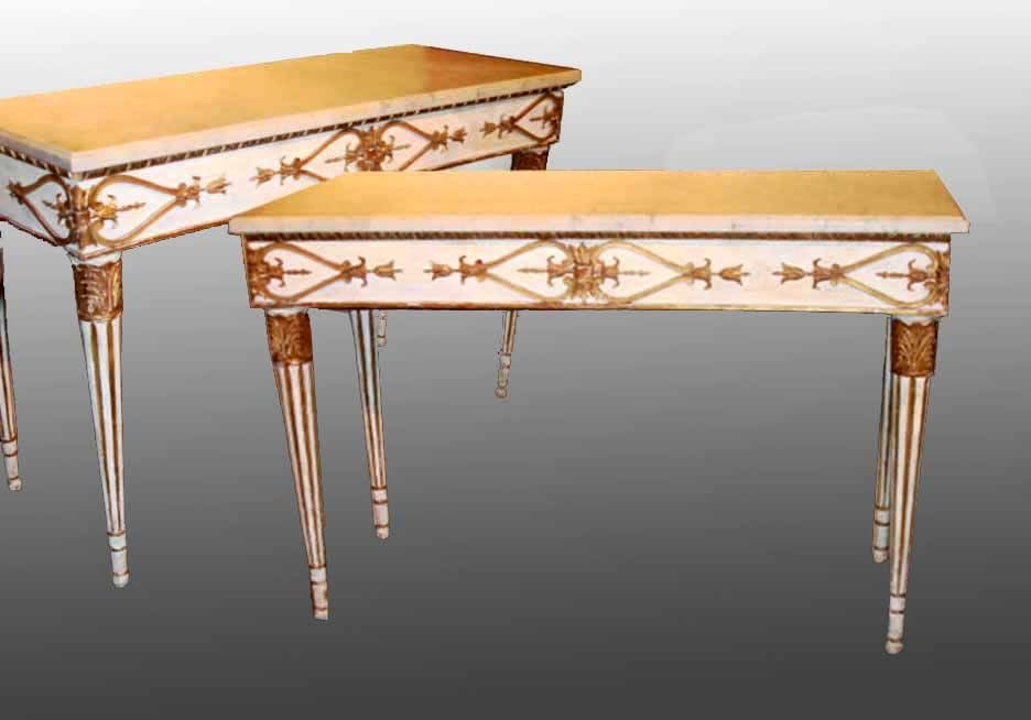 A fine PAIR of Italian neoclassical white and gold console tables. The bone white marble tops are above apron with an antique white background finely carved with classical rosettes and bell flowers and stylized palmettes. The tables are raised on