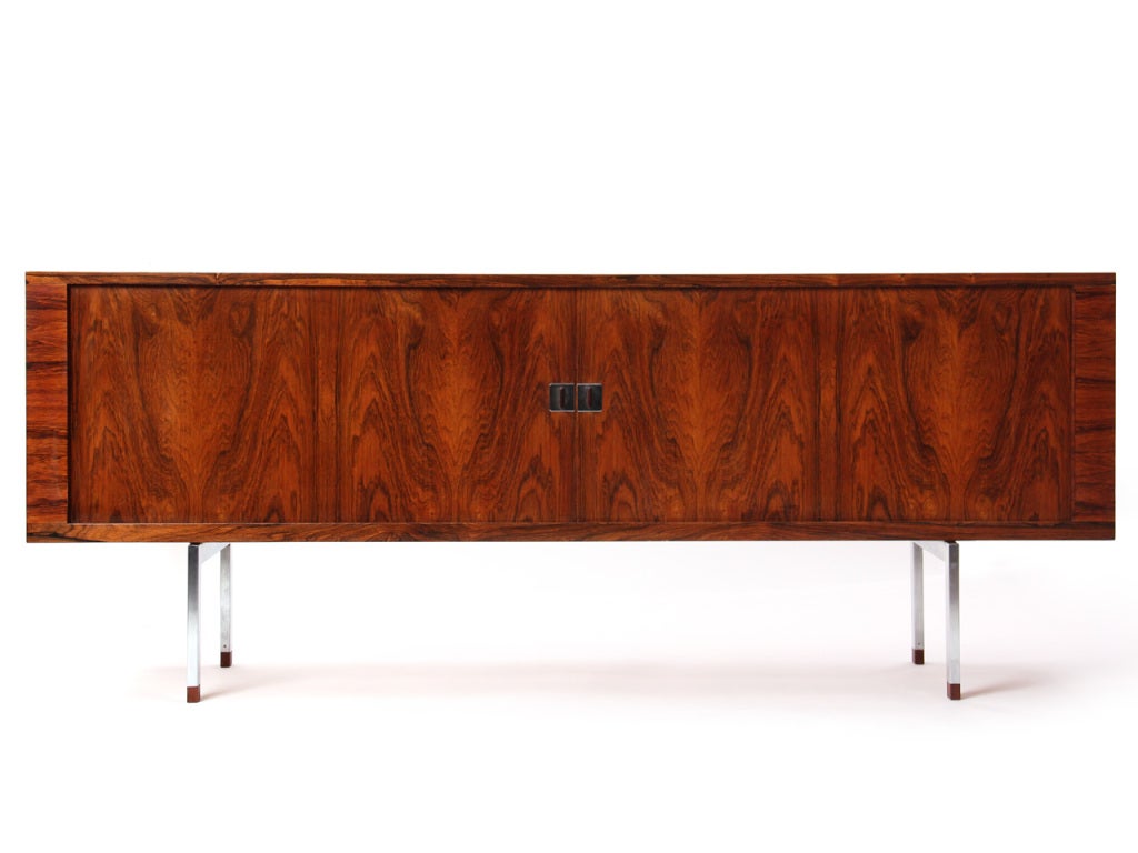 A rosewood credenza with two nearly invisible tambour sliding doors, enclosed, four shelves, and two adjustable trays on square chrome legs with rosewood feet.