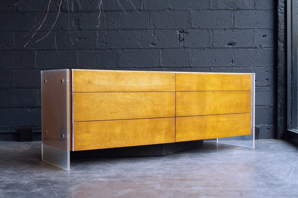 more commonly seen with wood side panels & full width plinth base, this version has lucite panels and a triangular plinth base; grain of figured wood continuous across 6 drawers; polished aluminum trim; retailed by John Stuart in New York City