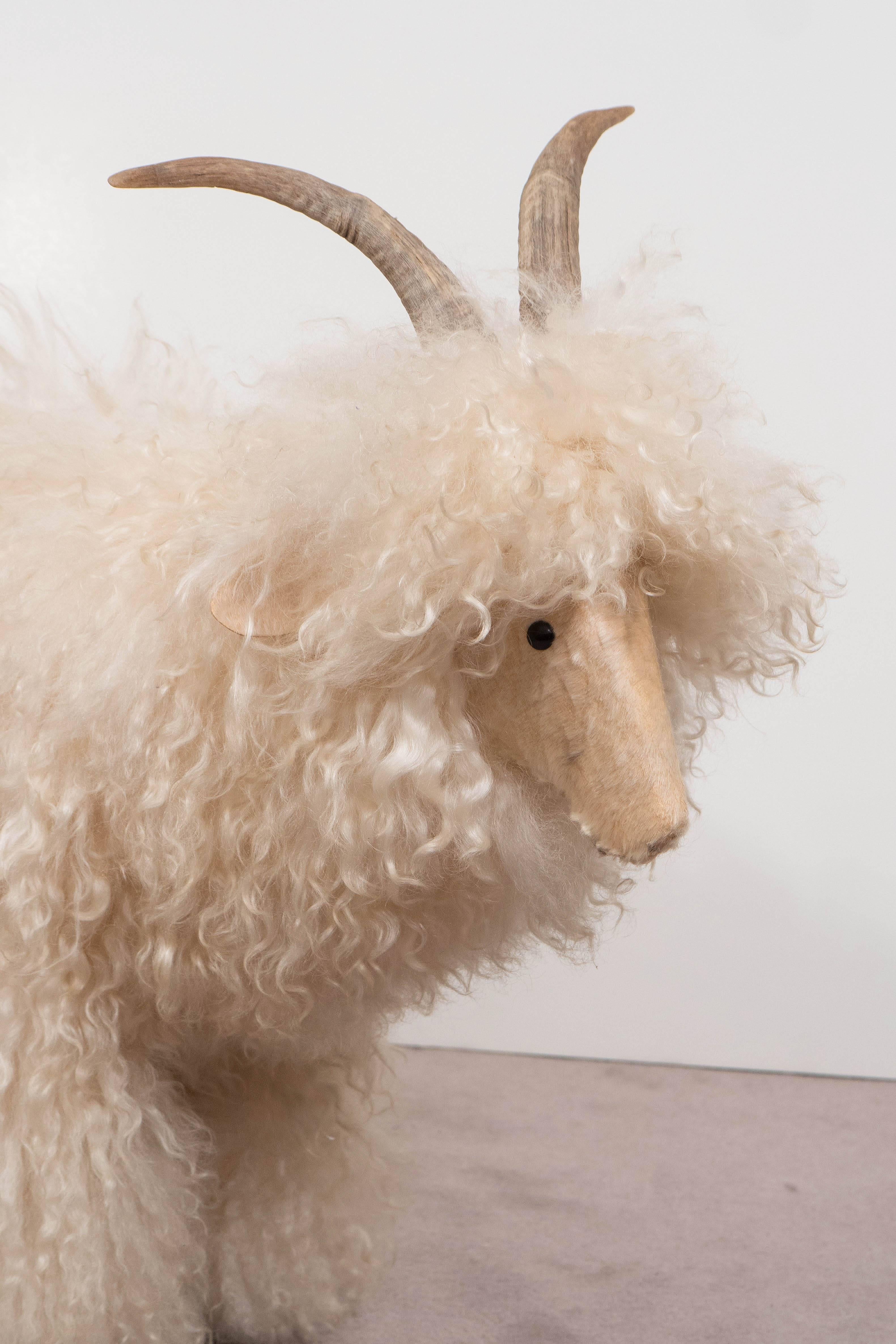 A sculpture of a white sheep, featuring genuine sheepskin and horns, with wooden legs and black buttons for eyes. It is reminiscent of the style of Francois Xavier Lalanne, French artist known for his ‘Herds of Sheep’ created in the 1960s. The