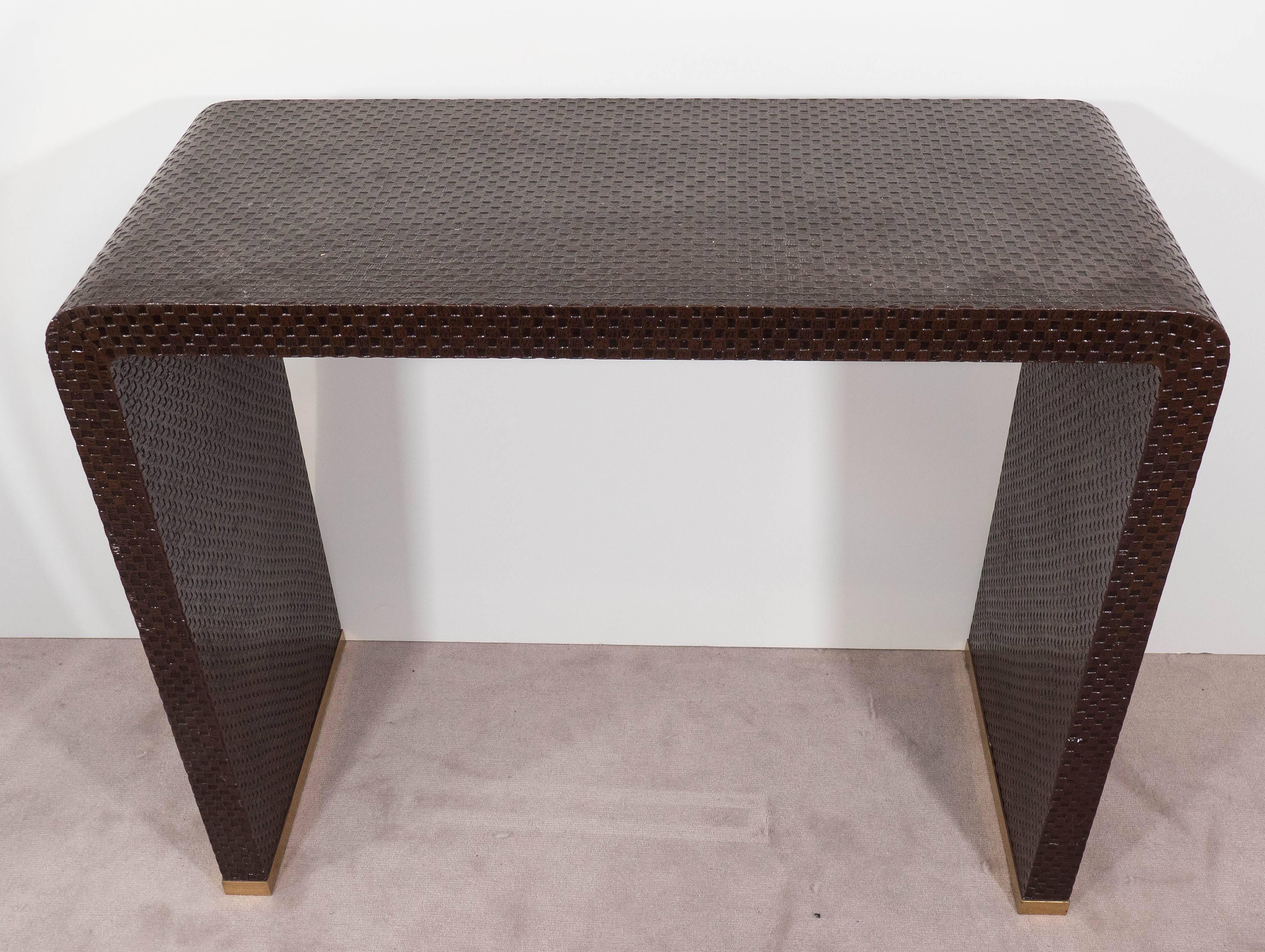 A waterfall console table, produced circa 1970s, with woven and lacquered grasscloth surface, against wood, with brass runners around the base. Good vintage condition, consistent with age and use.