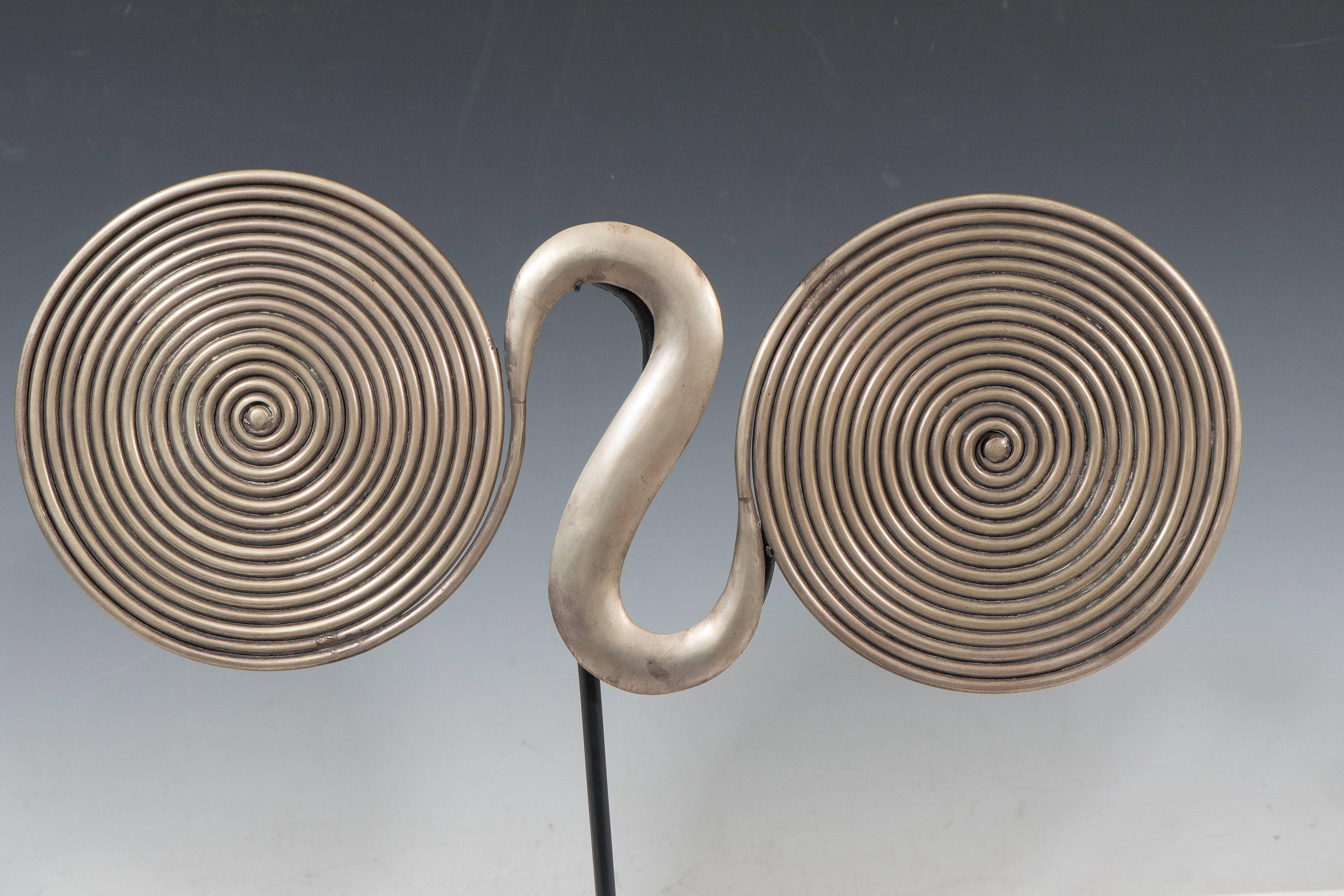 A vintage brass sculpture, influenced by Miao Hook tribal talismans of China. It consists of two tightly wound coils connected with a stylized hook, set on a black metal stand. Good vintage condition.