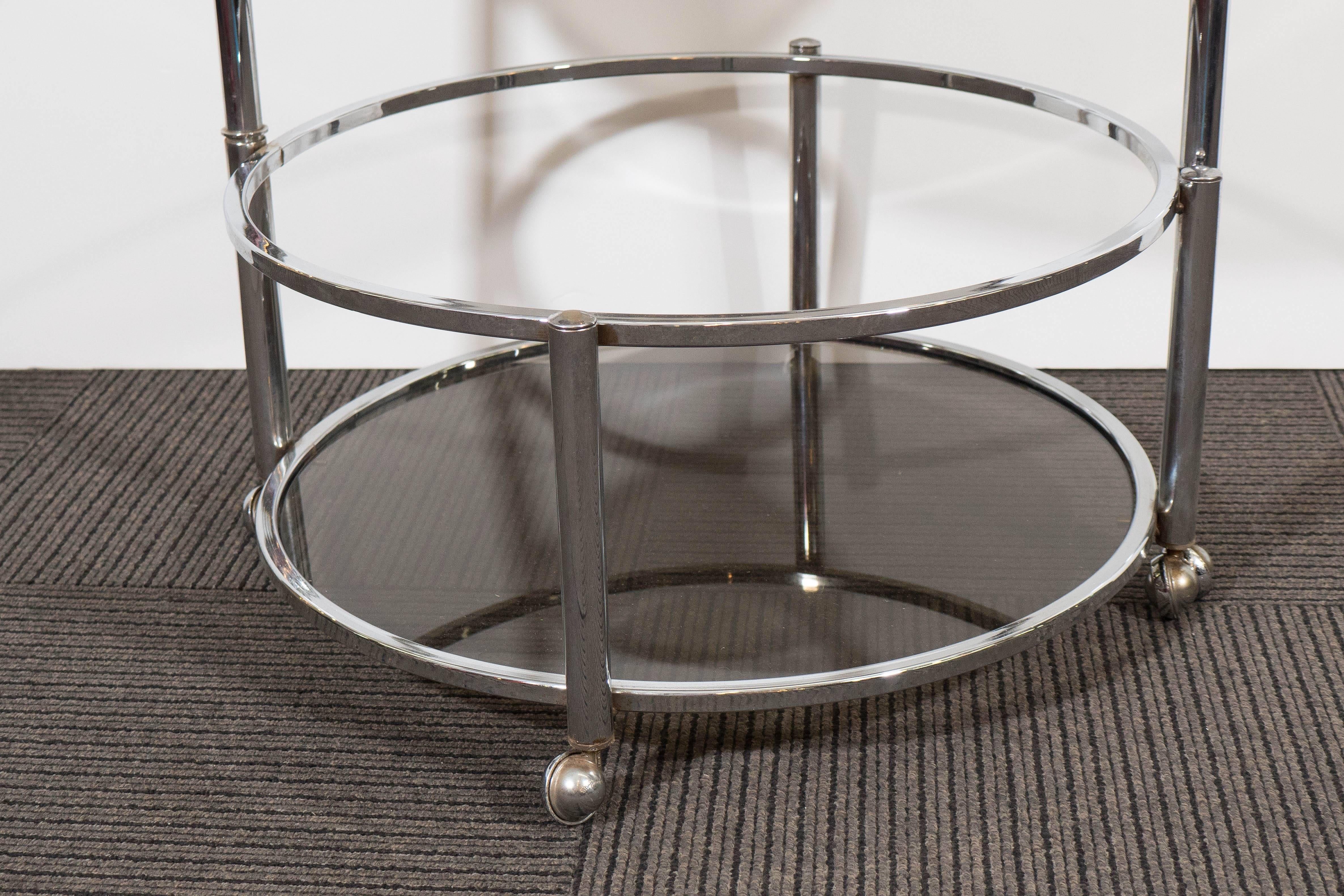A vintage circa 1970s round occasional table, with two shelves of smoked glass tiers, against an adjustable, beautifully polished chrome frame on casters; the central ring acts as a handle, permitting the bottom shelf to swivel outwards. Very good