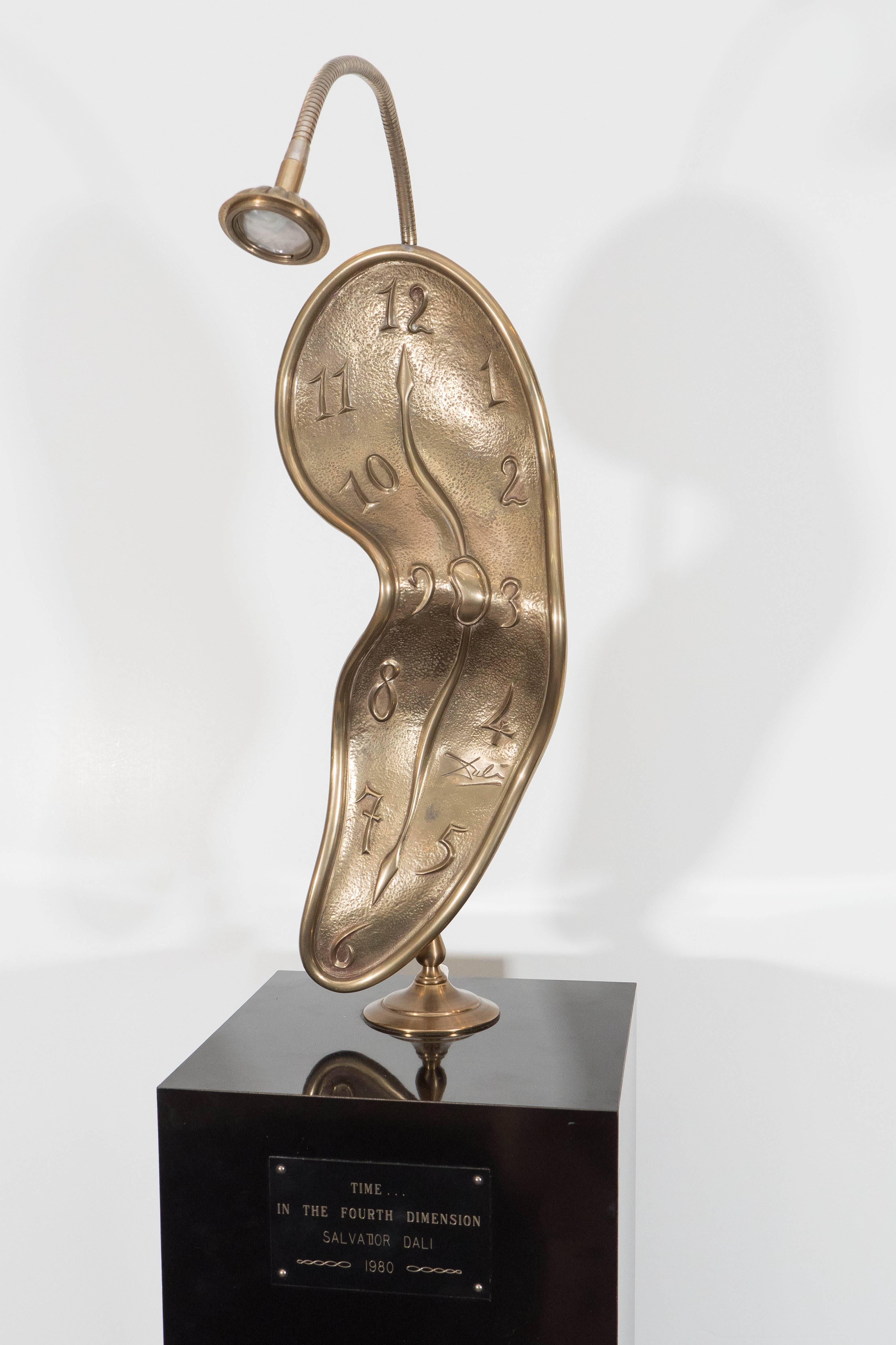 Dali's iconic melting clock image comes alive in sculptural form. Done in brass and mounted on a black acrylic pedestal, the piece is also wired to be a floor lamp with a movable pin bulb affixed above and a switch on the back. Plaque on front reads