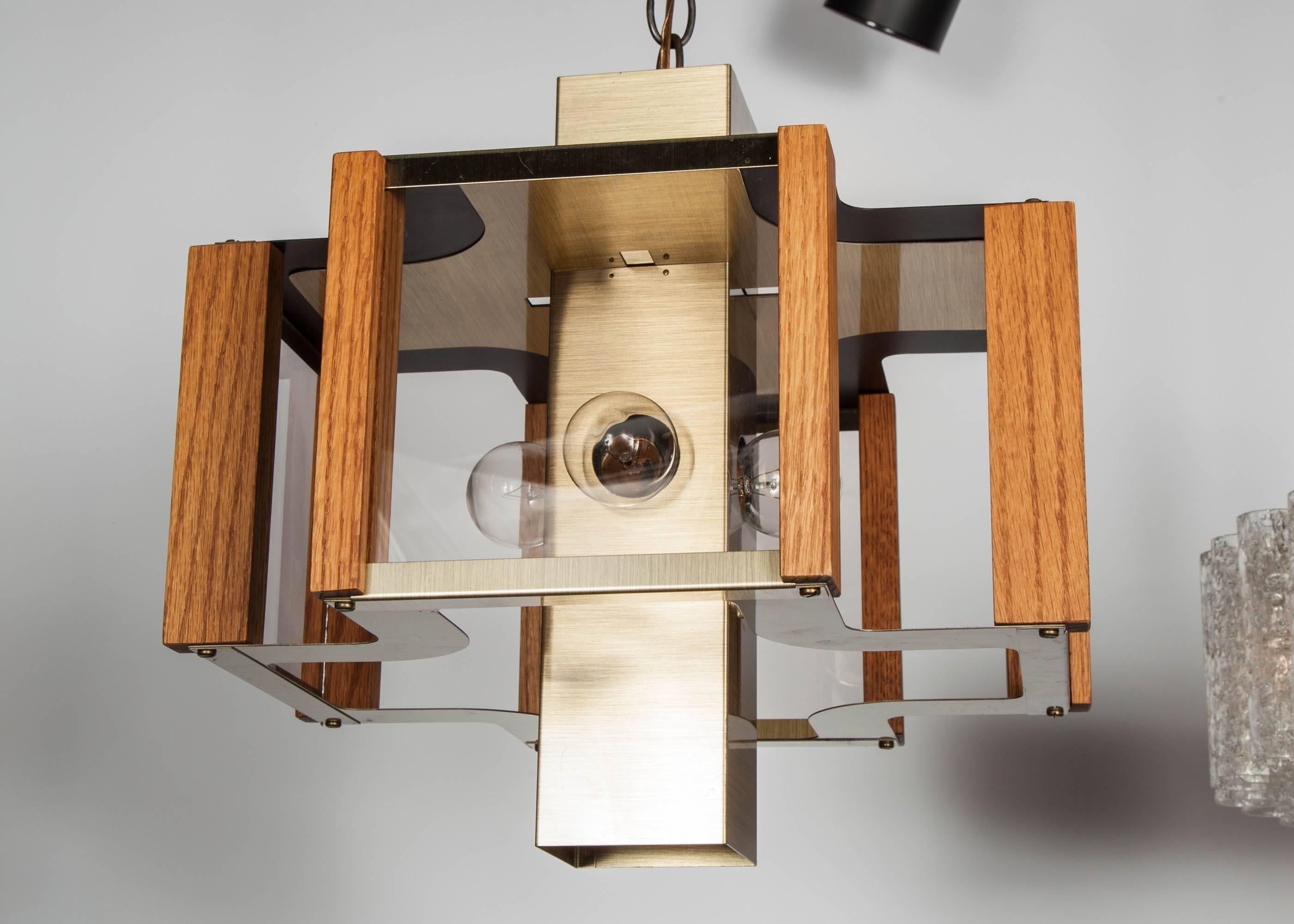 Modernist chandelier with sculptural design. The light fixture features an quadrilateral form comprised of a brushed brass metal frame with walnut wood trim details and smoked glass panels. Fixture has a square column center stem with four equal