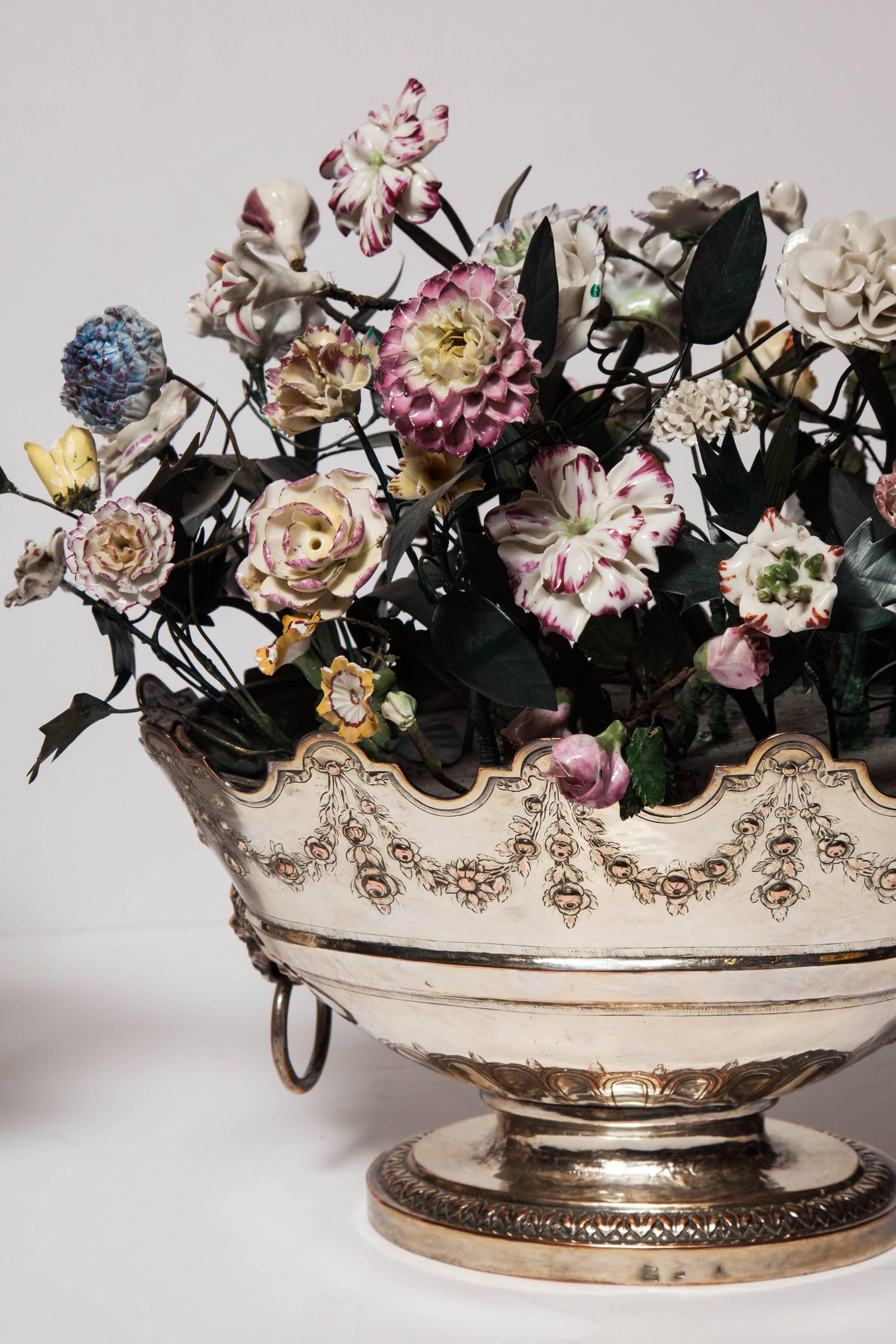 A fine pair of antique Louis XVI neoclassical style, tole piente, bronze and porcelain flowers finely mounted in a pair of oval shaped silvered bronze baskets (Jardeniers), each sculpted with a pair of lions masks holding a ring as handles connected