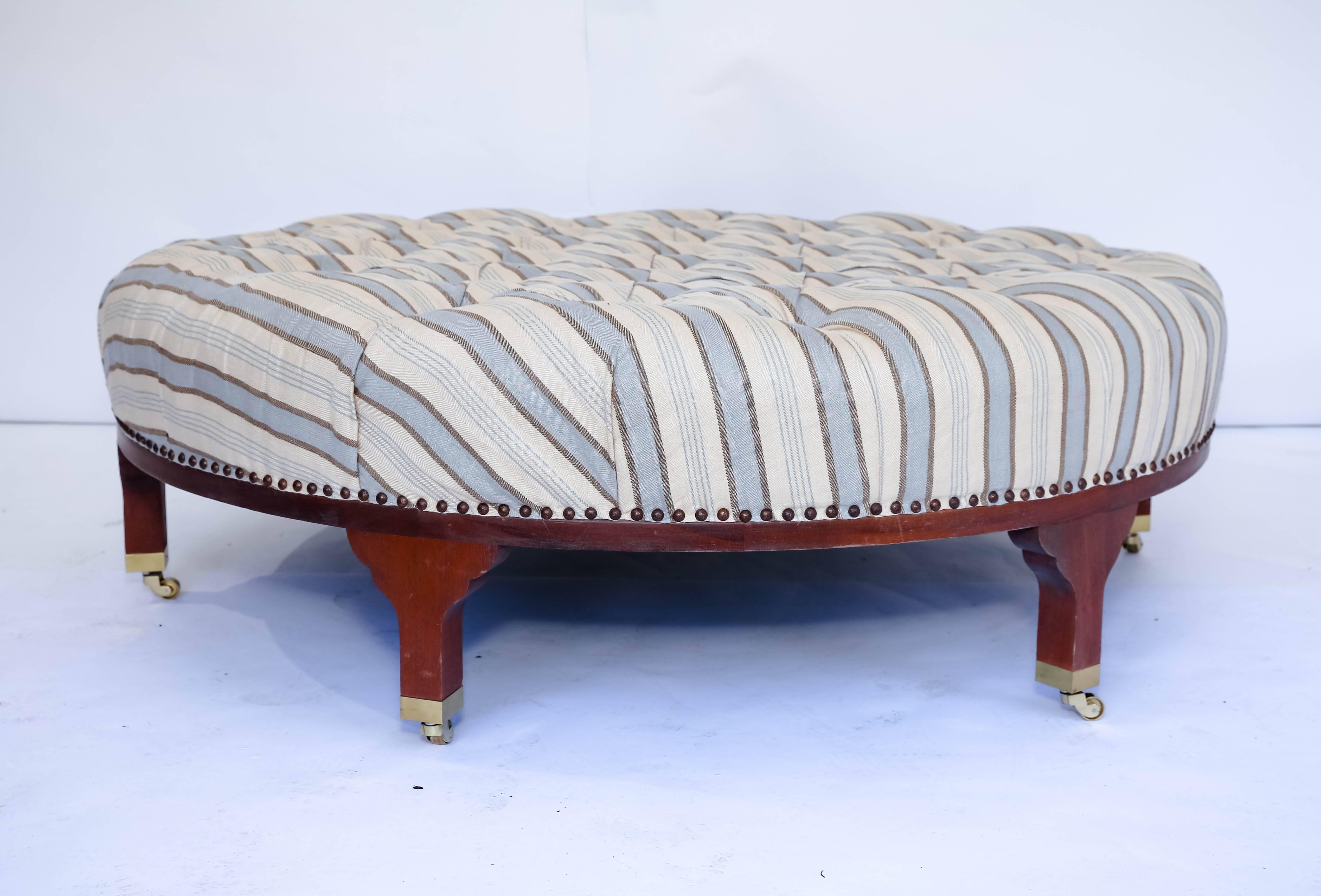 Round tufted ottoman or coffee table in striped linen upholstery. Wooden base set upon brass casters.   

