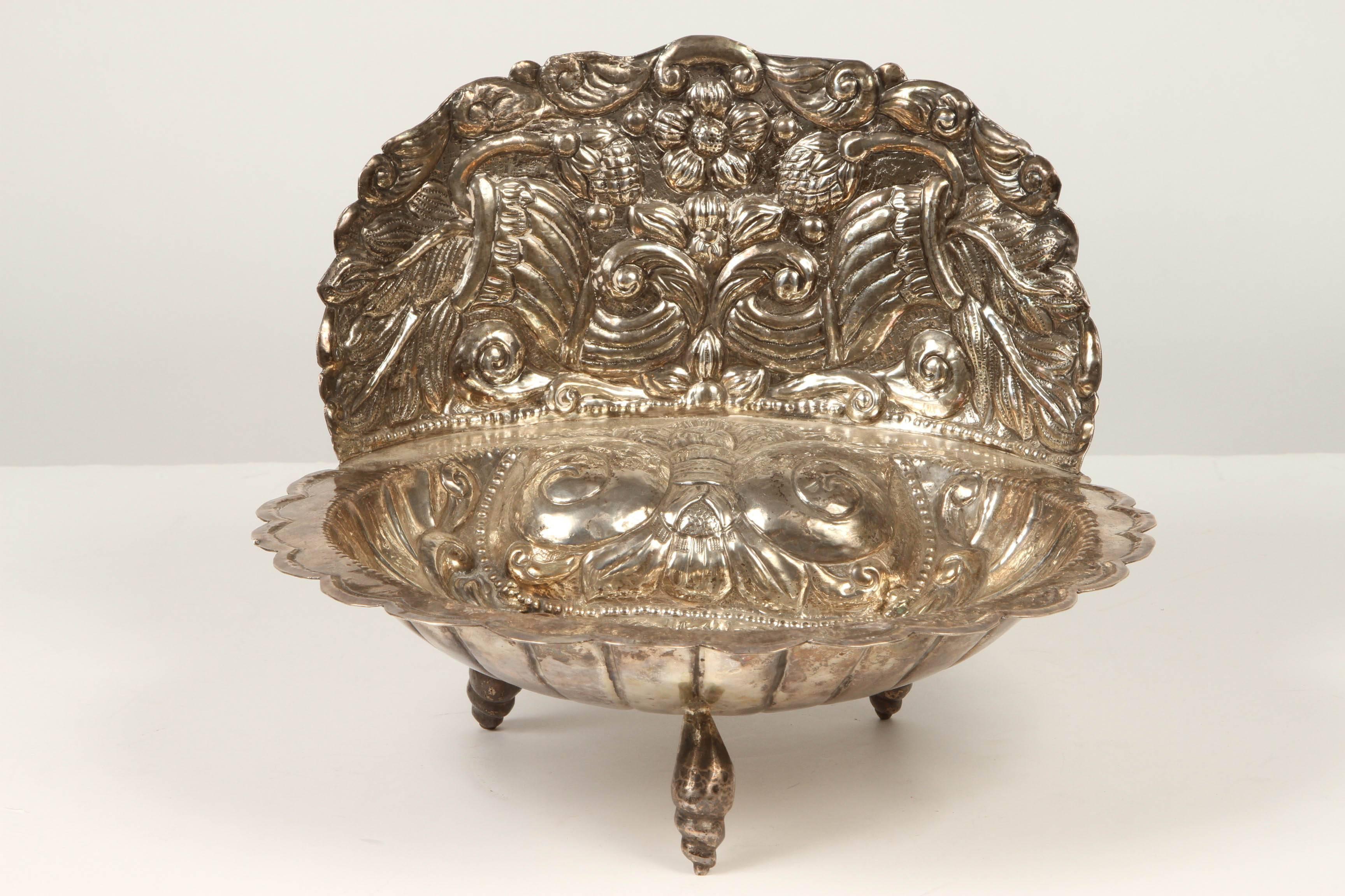 A silver font, covered with scrolling and floral patterns in the silver. Four feet and a raised sort of backsplash which is also elaborately engraved.