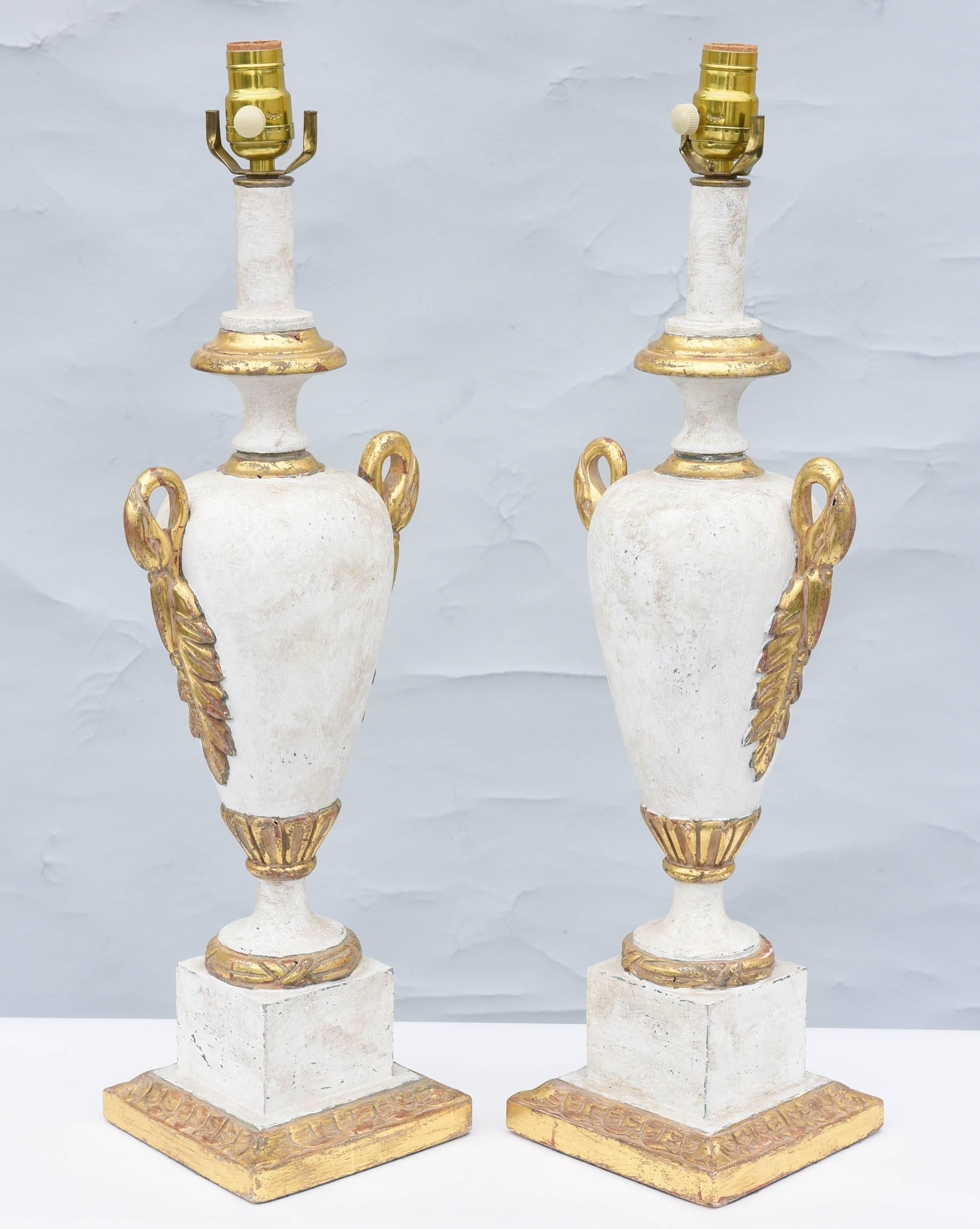 Pair of painted and parcel gilt lamps, each a wooden urn form, with white, mottled, and gessoed finish, flanked by gilded swan neck and acanthus handles, on round foot and square plinth.  

Stock ID: D7344