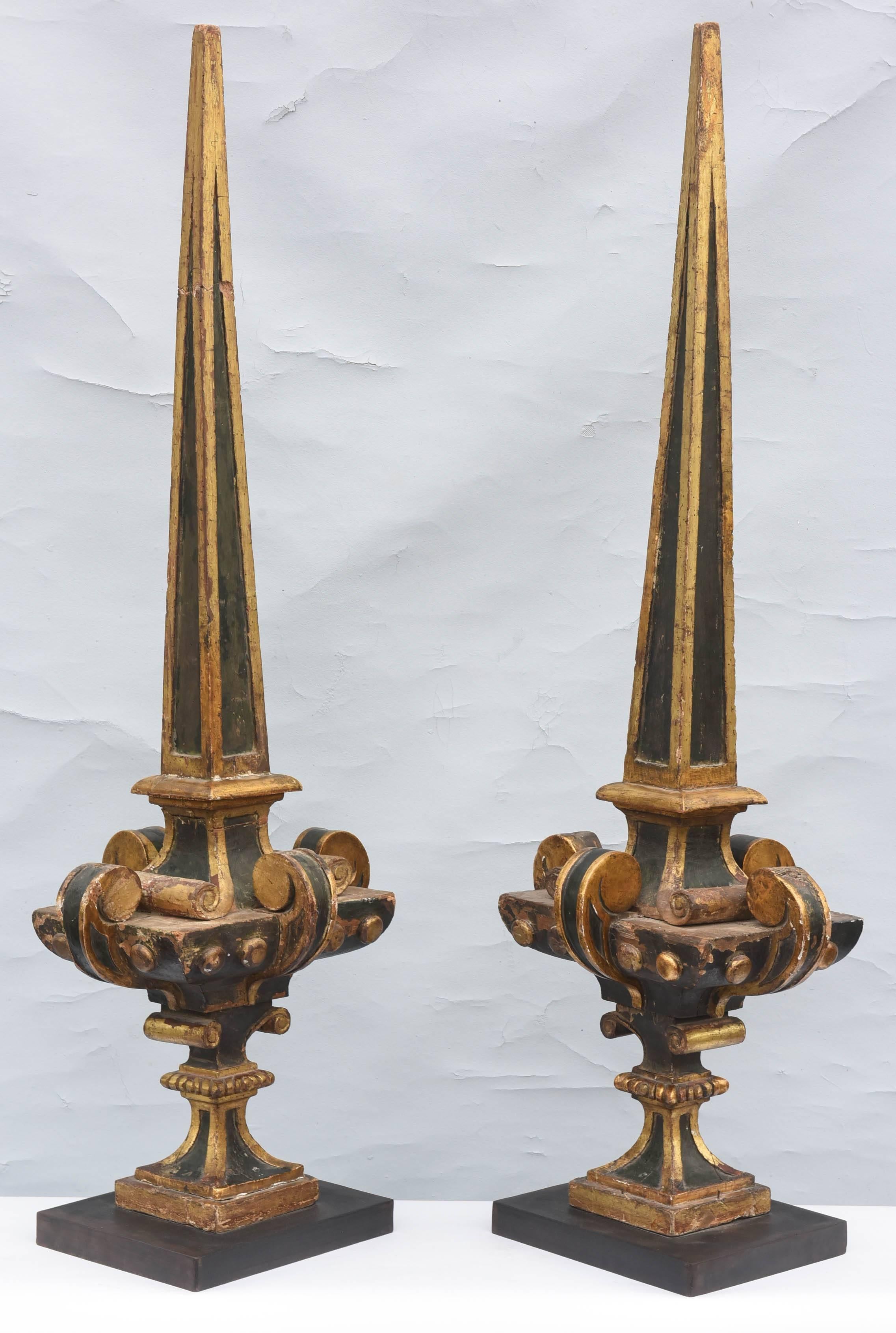 Monumental pair of finials, black-painted and parcel-gilt, of carved wood, each fielded obelisk spire, on urn-form base with scrolling handles, on beaded socle, surmounted by ionic capital, on square foot, mounted on black iron plinth.

Iron base