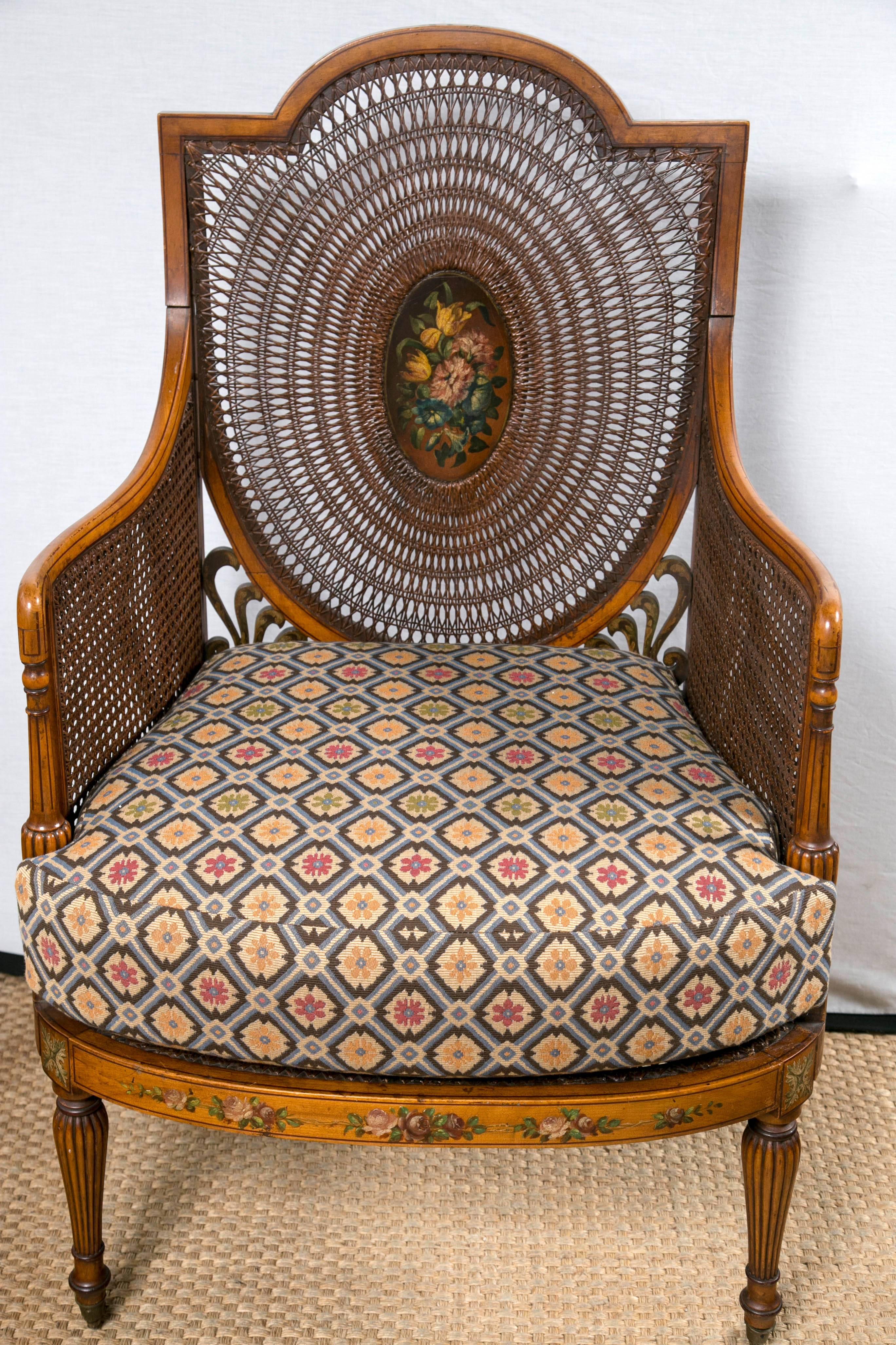 The arched top rail above a rounded caned back with inset painted oval plaque. The sides and seat fully caned. Round tapered and fluted legs in front and splayed legs in the back. Removable upholstered cushion. Arms, side rails and back painted in