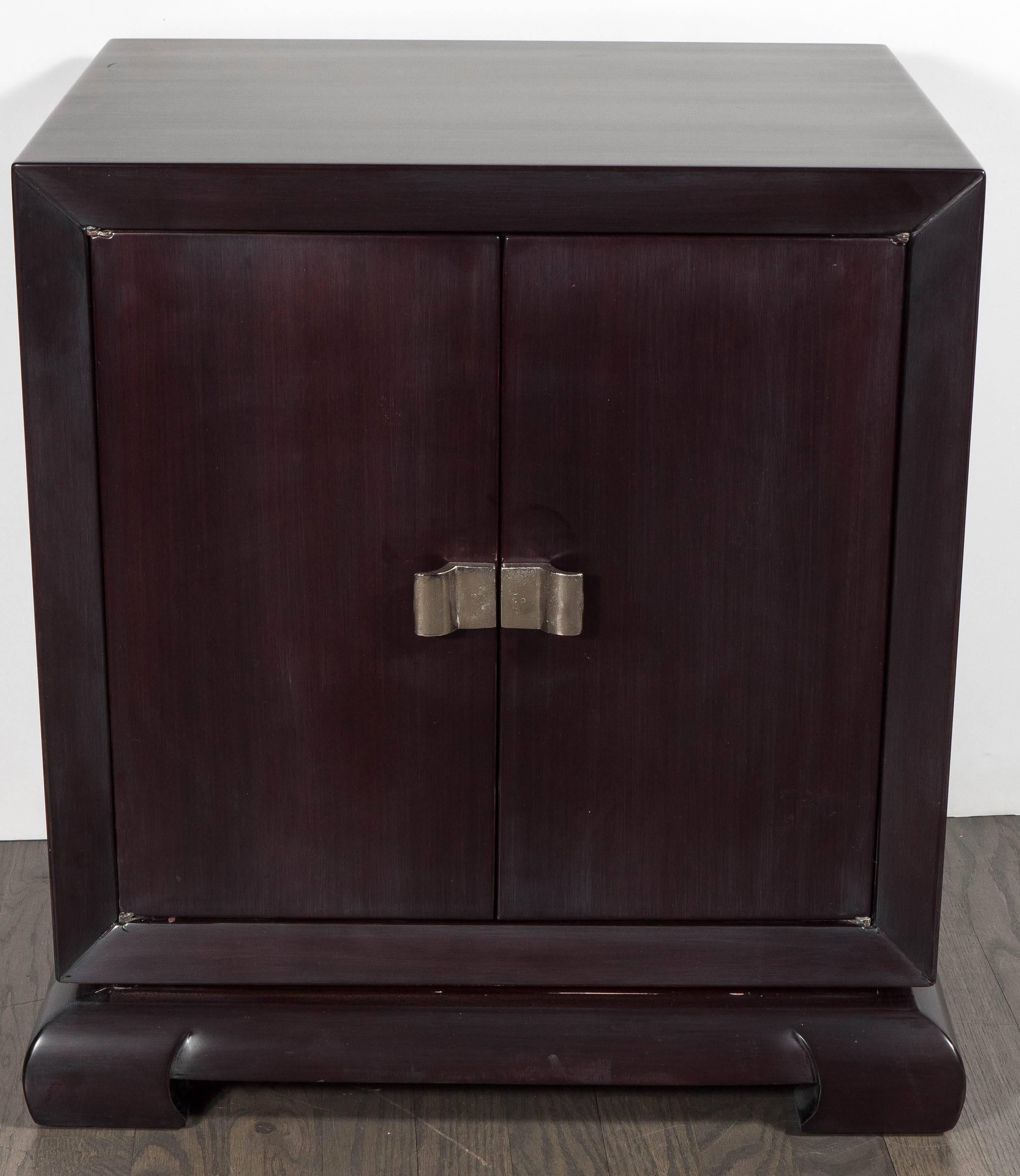This outstanding pair of Mid-Century Modernist nightstands or end tables in the manner of James Mont in hand rubbed rich brown mahogany feature a pair of center opening doors with horizontal stylized bowed design nickel pulls and stylized scroll