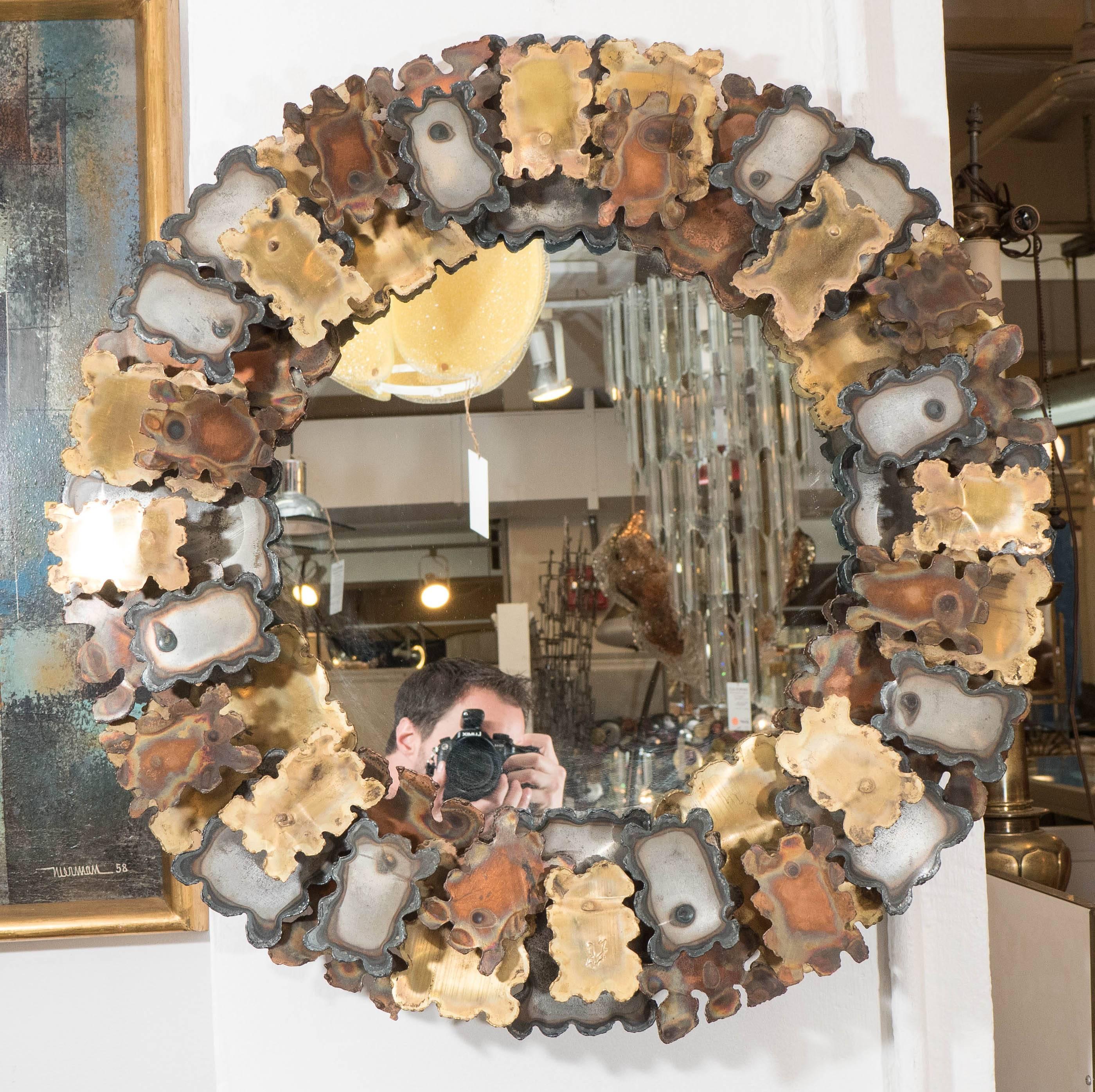 A round wall mirror, with an elaborate frame designed in the manner of Brutalist artist and designer Silas Seandel, decorated with rough and distressed cutouts of mixed metal sheets in three different tones, circa 1970s. Markings include signature,