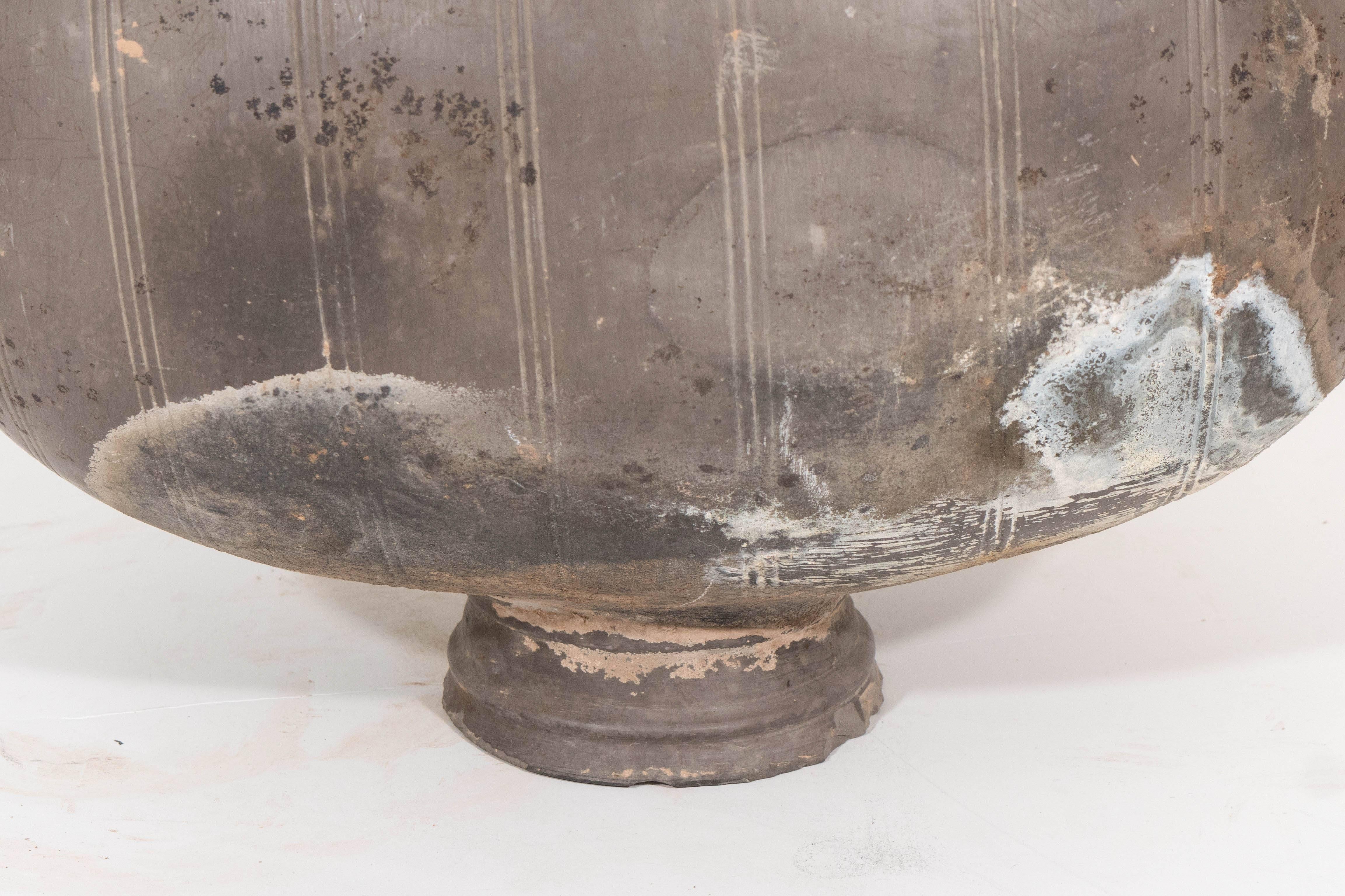 An ancient Chinese pottery 'cocoon' jar, produced within the Han Dynasty period, so-called for its semblance to a silkworm cocoon, with vertical incised lines surrounding the body, raised on a single foot. Other than some spotting and wear,