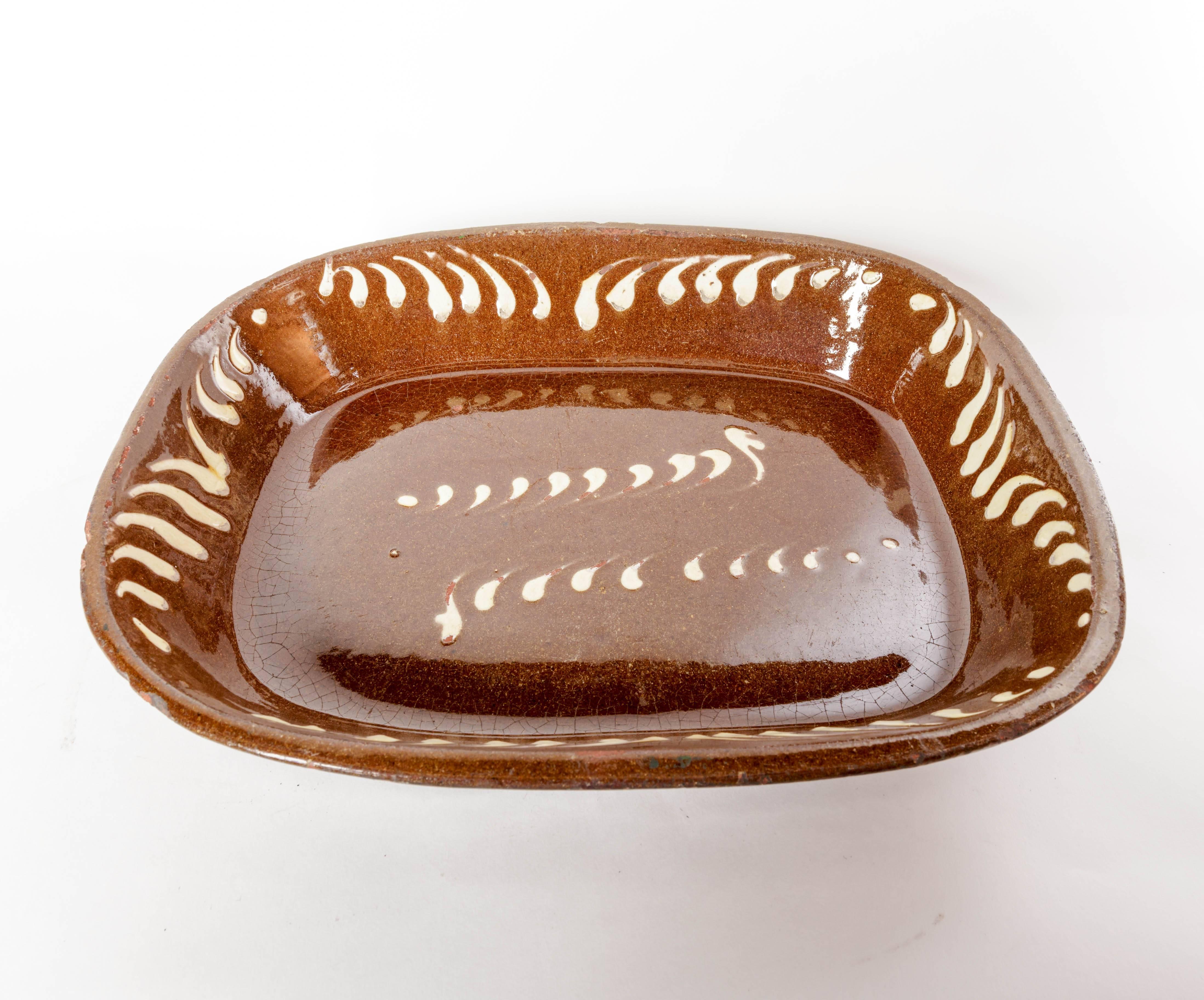Large 19th century English slipware baking dish with off-white decoration. The tapered sides meet to form a rounded rectangular base. The gloss brown glaze extends down two thirds of the exterior side. The off-white squiggle decoration is on all