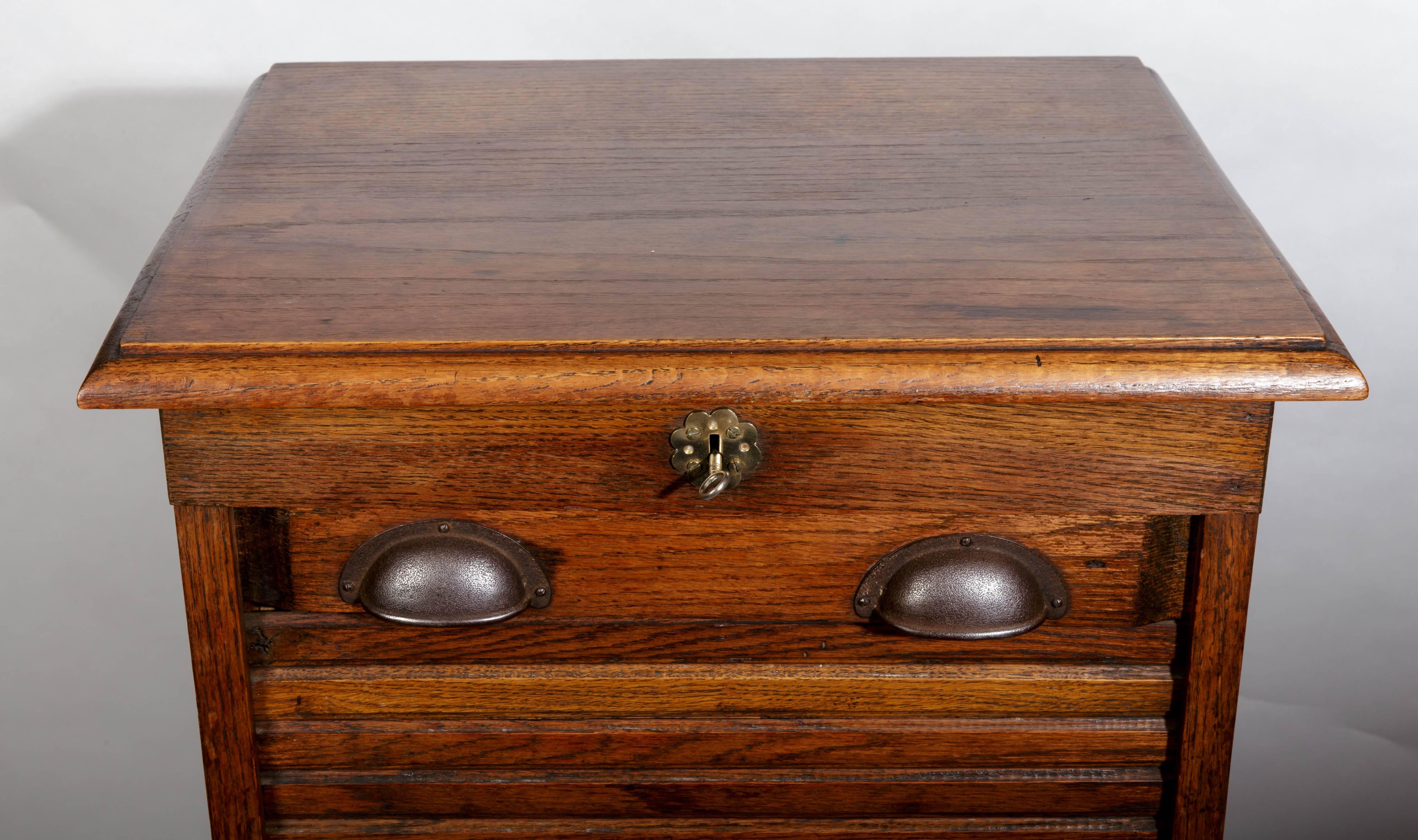 French oak tambour front (roll down) file cabinet. The tambour front is locked by a key at the top center having a floral shaped brass escutcheon plate. A pair of burnished iron handles allow the lowering and raising of tambour panel. The interior
