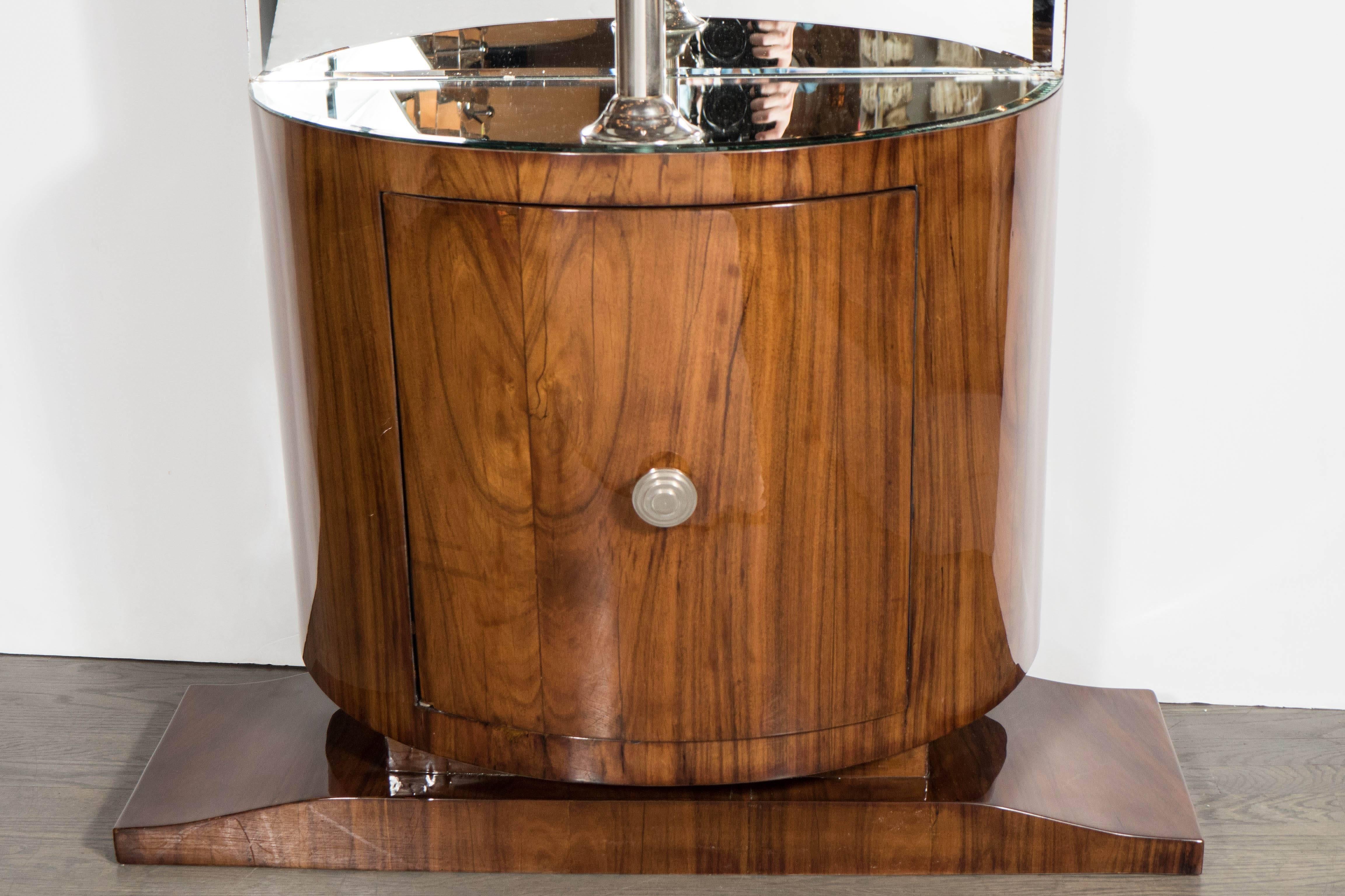This exceptional streamlined Art Deco demilune bar in fine bookmatched walnut sits atop a tapered plinth base. It has a cabinet door with a single skyscraper style nickeled pull beneath a mirrored niche with a nickeled vertical support for
