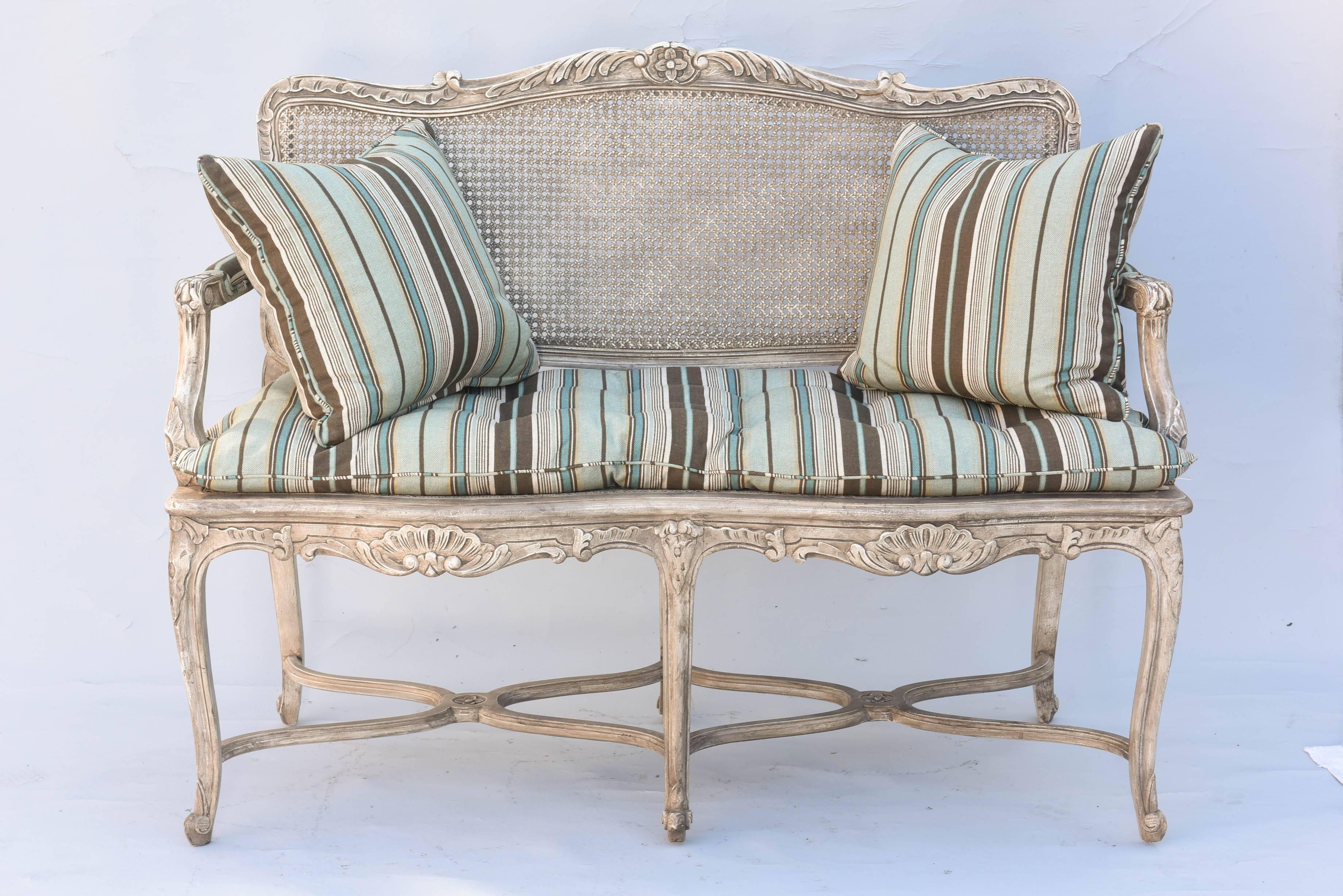 Settee, in Regence taste, having a painted frame, showing natural wear, its serpentine crest rail carved with a rosette, flanked by foliate ends, having a caned back and seat, with padded armrests, on downswept terminals, a loose cushion, on