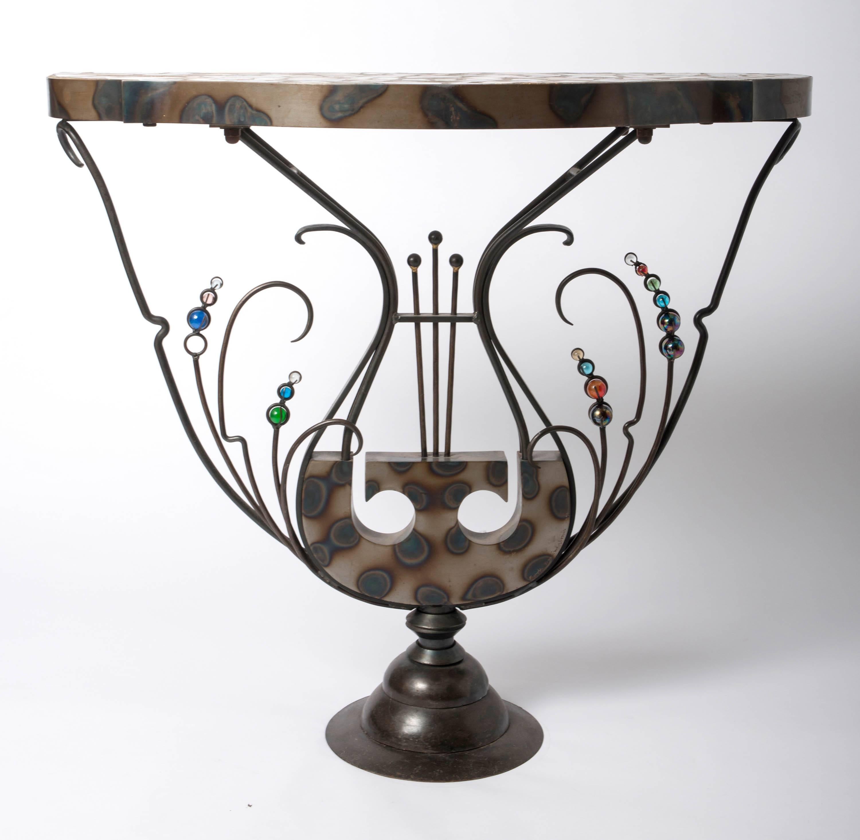 A “Lyra” console table, by Mark Brazier-Jones.
Forged iron, burnt metal, glass balls, various marble fragments embedded in cement.
Incised signature- “Mark Brazier-Jones 1991,”
England 1991
Measure: H 90 cms, W 103 cms, D 48 cms.
Provenance;