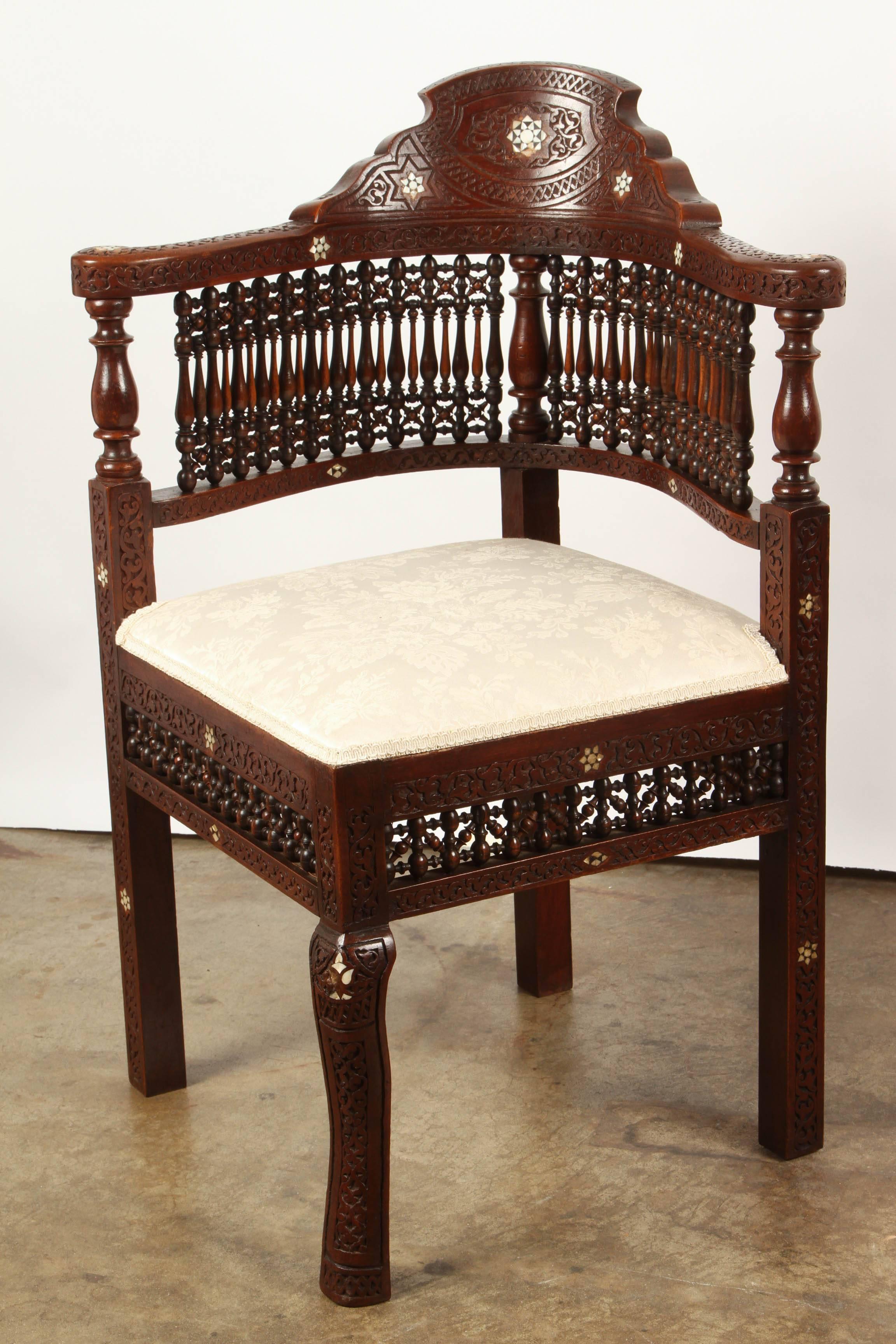 A pair of Moroccan spindle corner chairs, with carved and stained frames. The chairs have rails and arm rests, and the seats are upholstered in silk.The backs of the chairs are composed of a series of decorative spindles and are topped with a number
