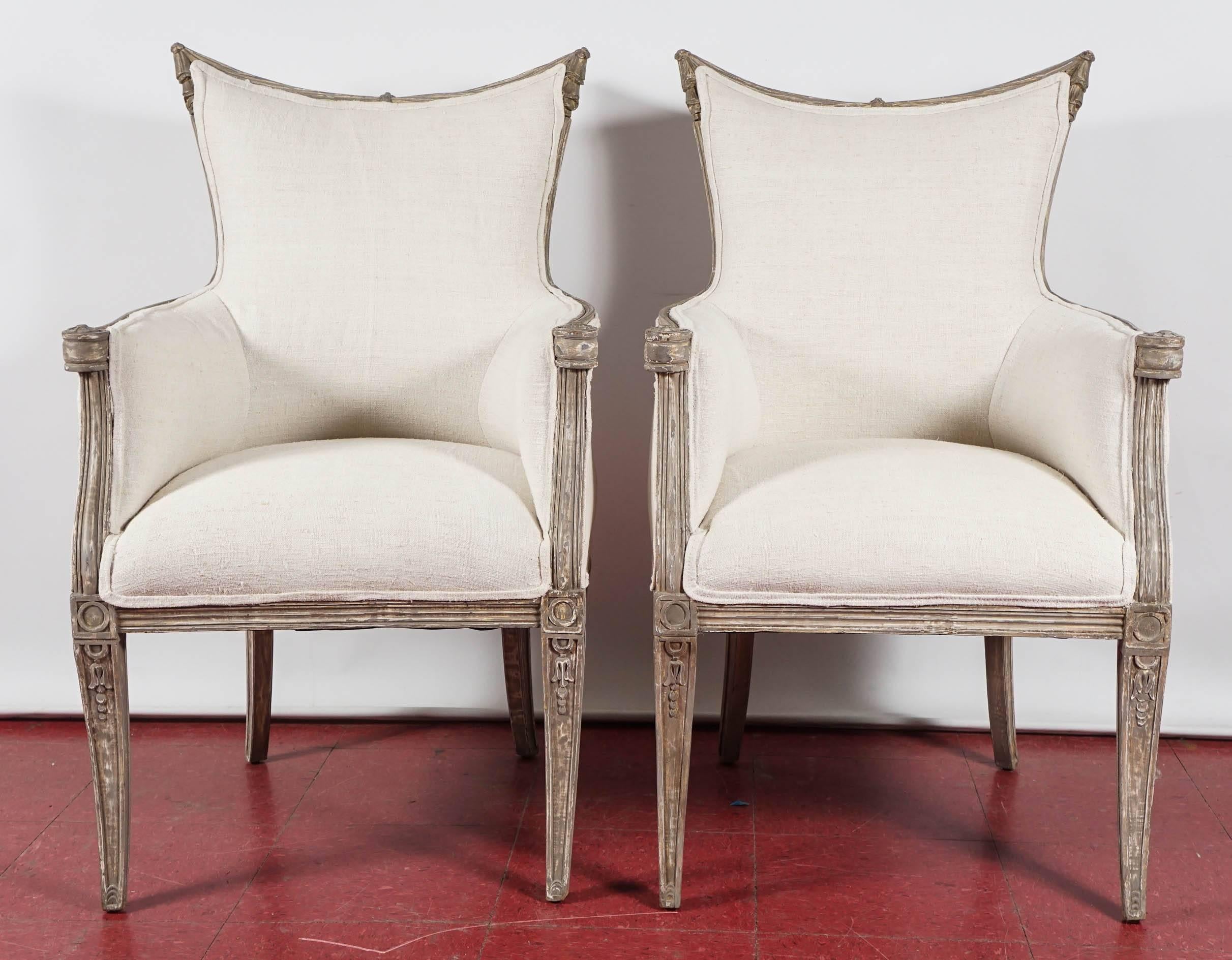 These Italian moderne burgers inspired by the Grosfeld House Hollywood Regency style are upholstered in beige linen and have hand-carved frames painted gray and are embellished with bell flowers carved within paneled front legs, while the ends of