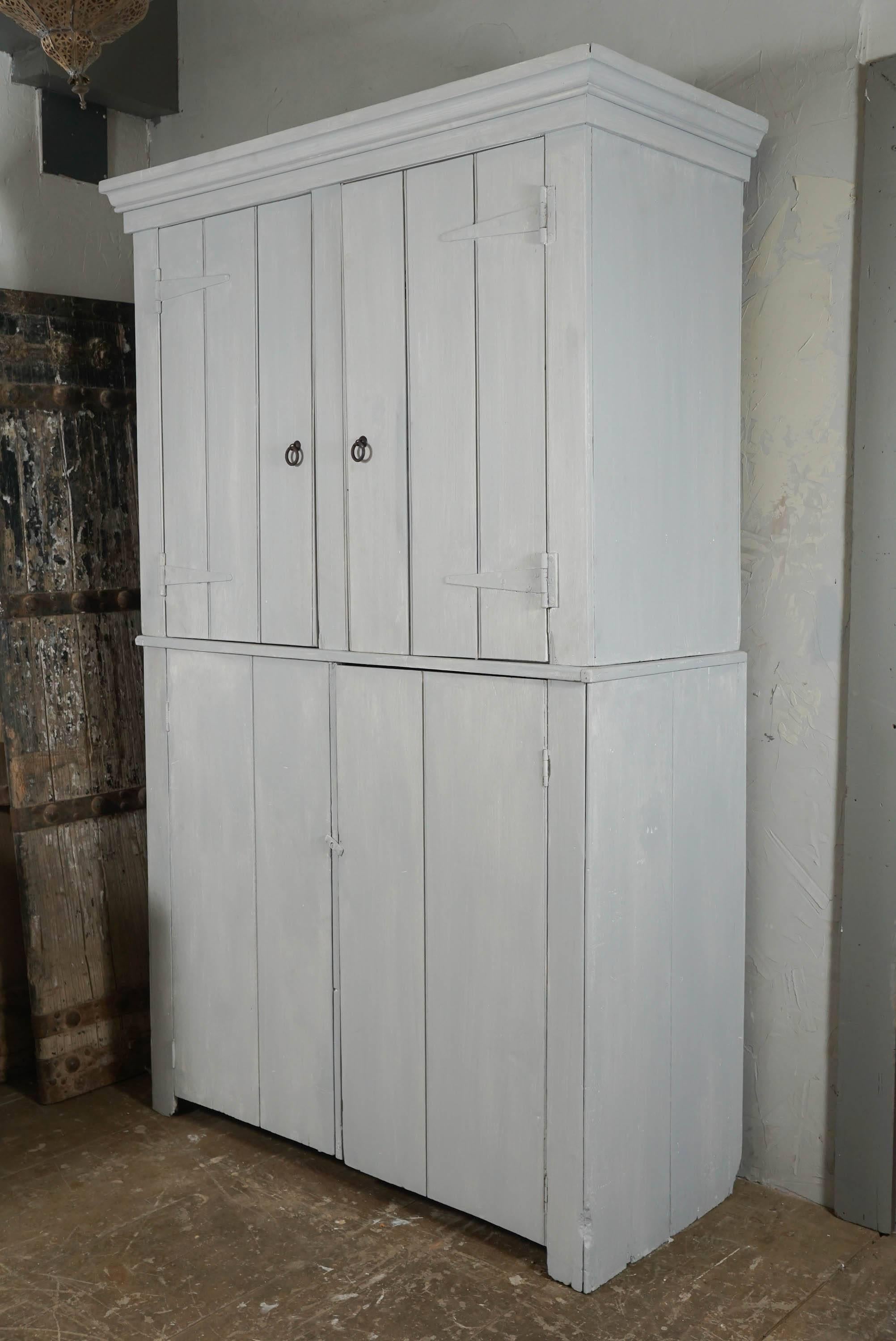 The capacious pale grey 19th century cupboard is constructed of separate top and bottom parts. The upper part has paneled double doors with wrought iron ring pulls and decorative hinges. Inside are two full shelves alternating with two half-depth