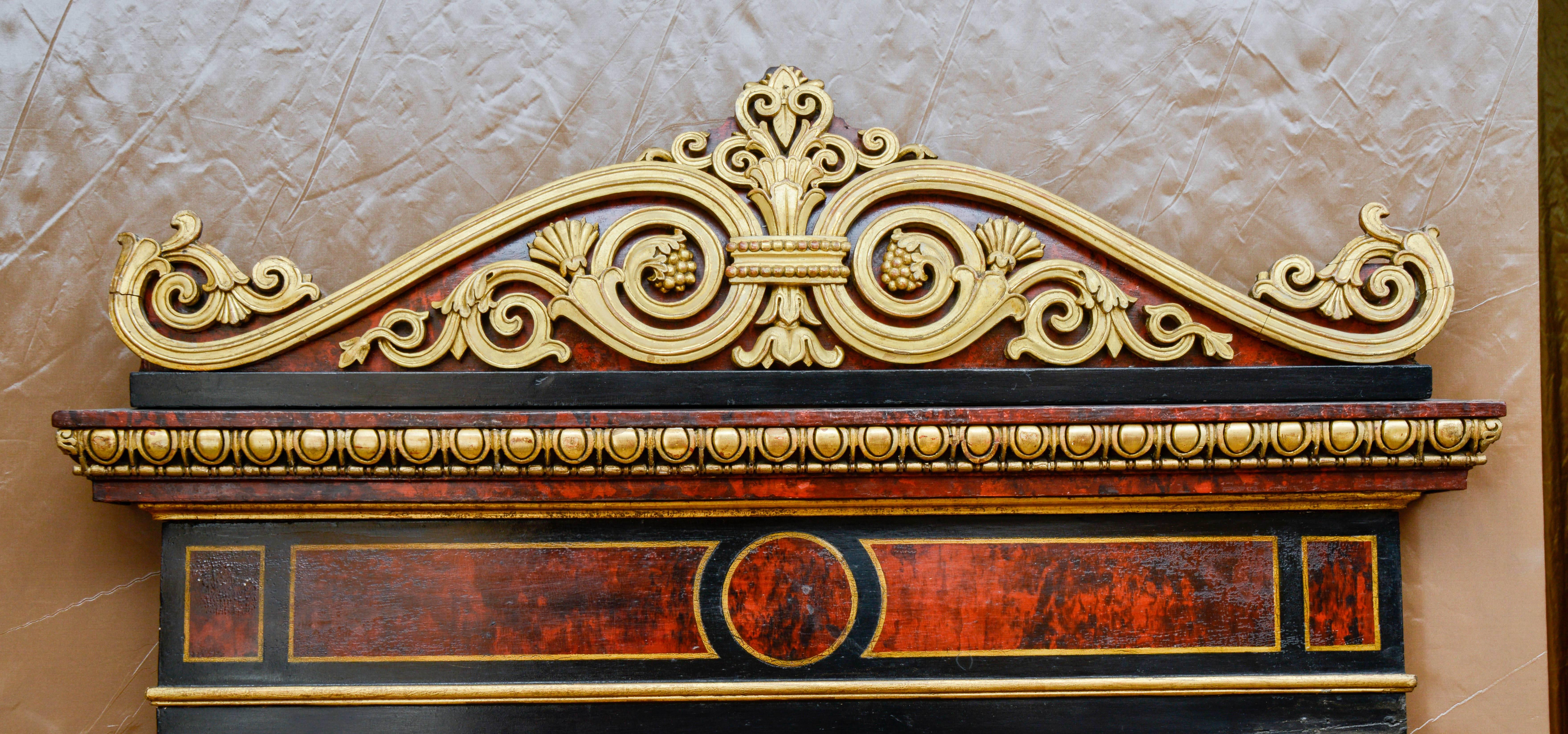 Trumeau Mirror in gilt and painted in black and red marble imitation wood, containing architectural motifs. It is framed by two Tuscan pilasters, surmounted by an entablature and a crown molding with modillions. The motif of the pediment is replaced