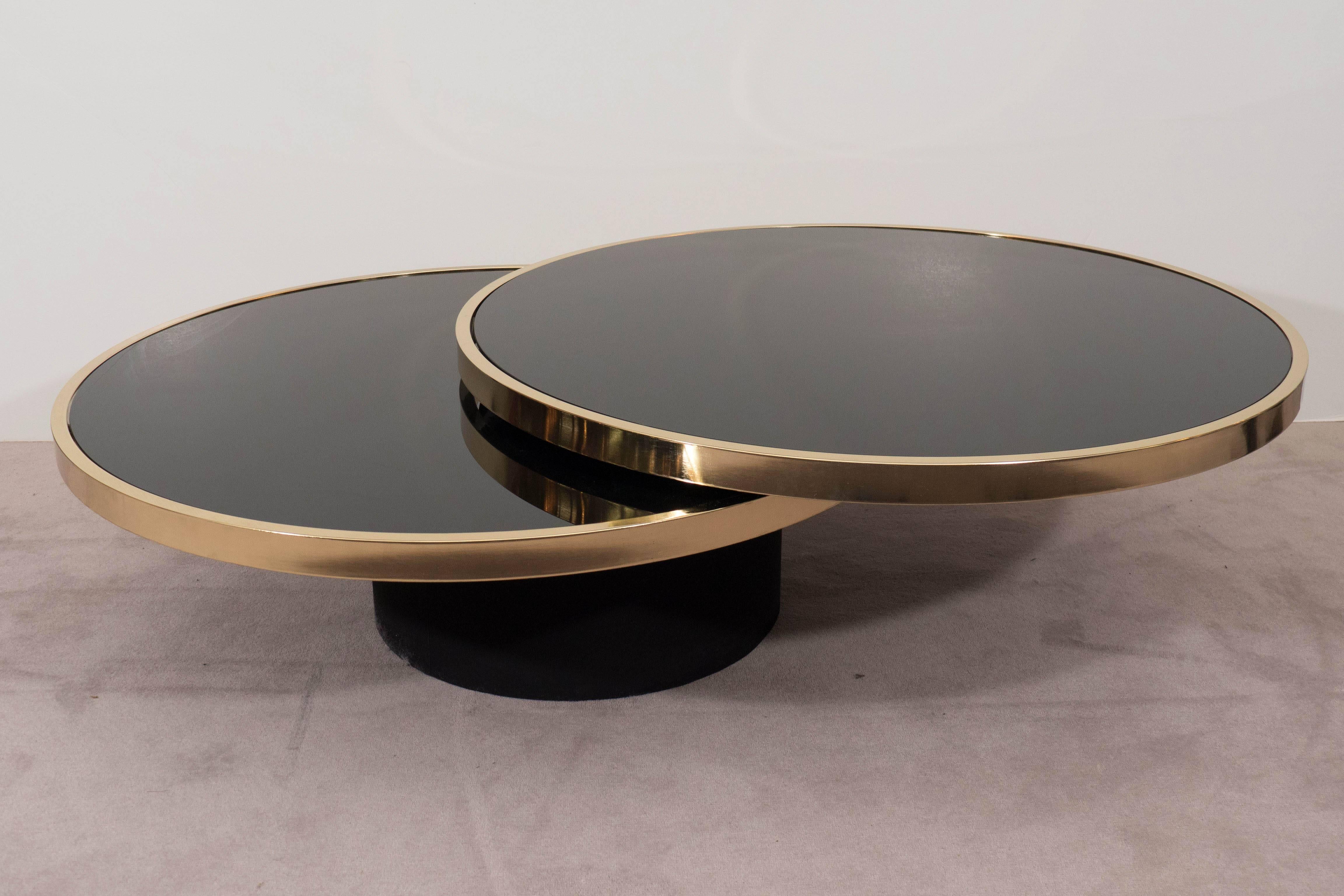 A vintage two-tier cocktail table, produced circa 1970s by the Design Institute of America (DIA), with gleaming black glass tops, in round brass frames, on a black cylindrical base, which enables the top tier to swing outwards. Fully extended the
