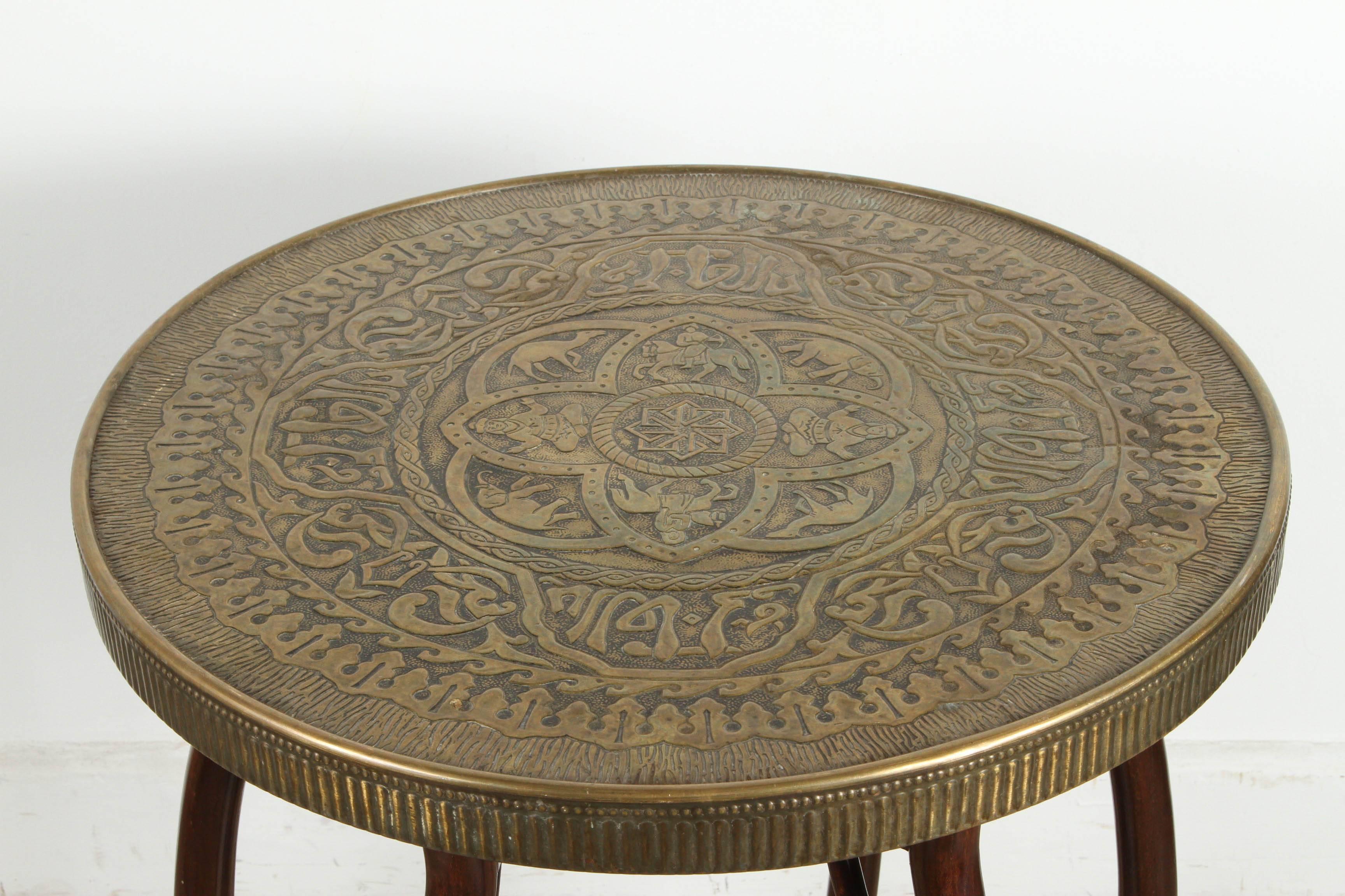 Middle Eastern brass tray table with bamboo stand, the top brass embossed top is decorated with calligraphy Arabic writing and figures of humans and animals in the middle medallion.
The top is removable and consist of a round wooden top covered