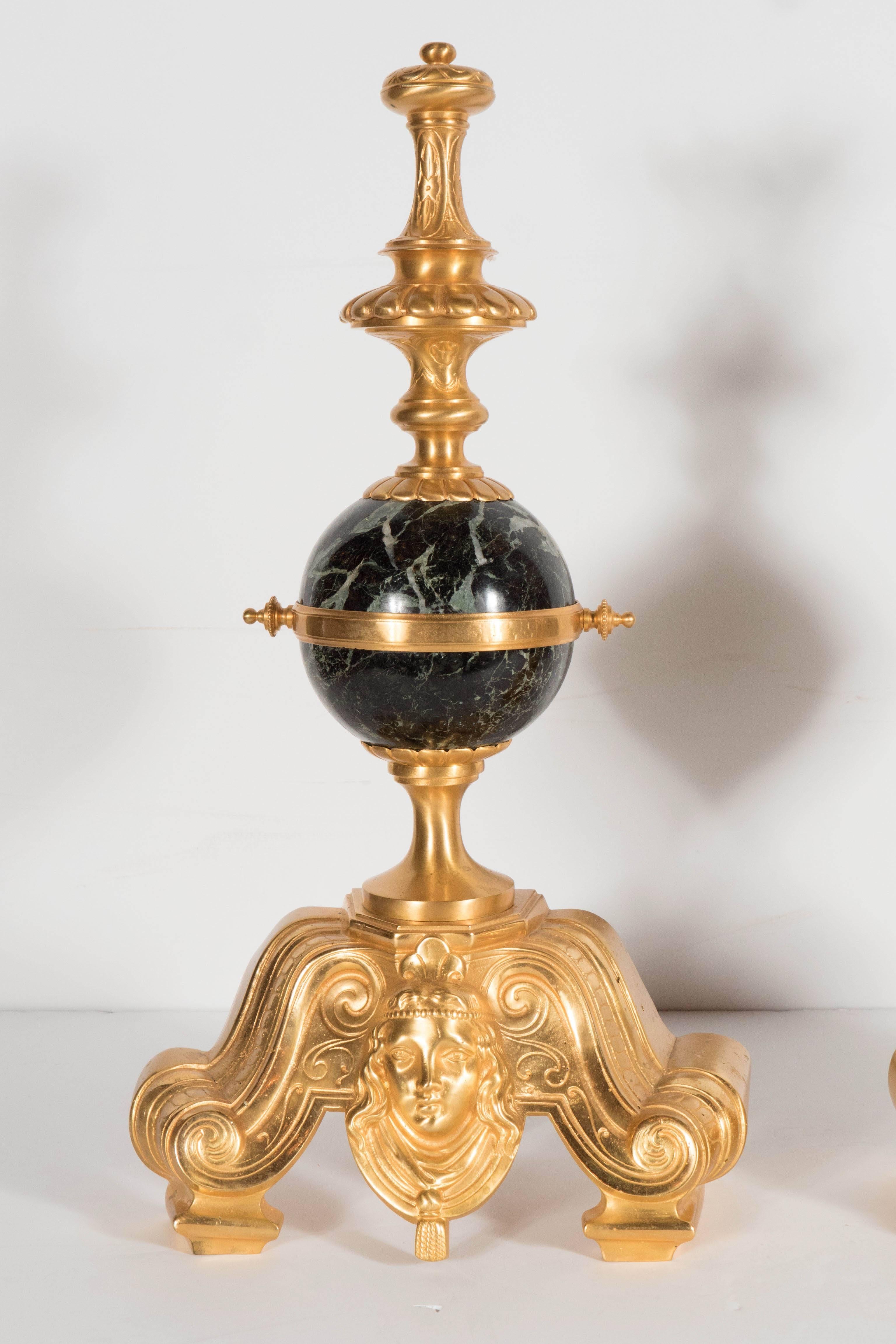 These stunning antique andirons chenets feature a Louis XV design in gilded bronze. A scroll form base with a bust adornment supporting a spherical exotic green marble detail with a stylized acanthus finial on top. These are in excellent
