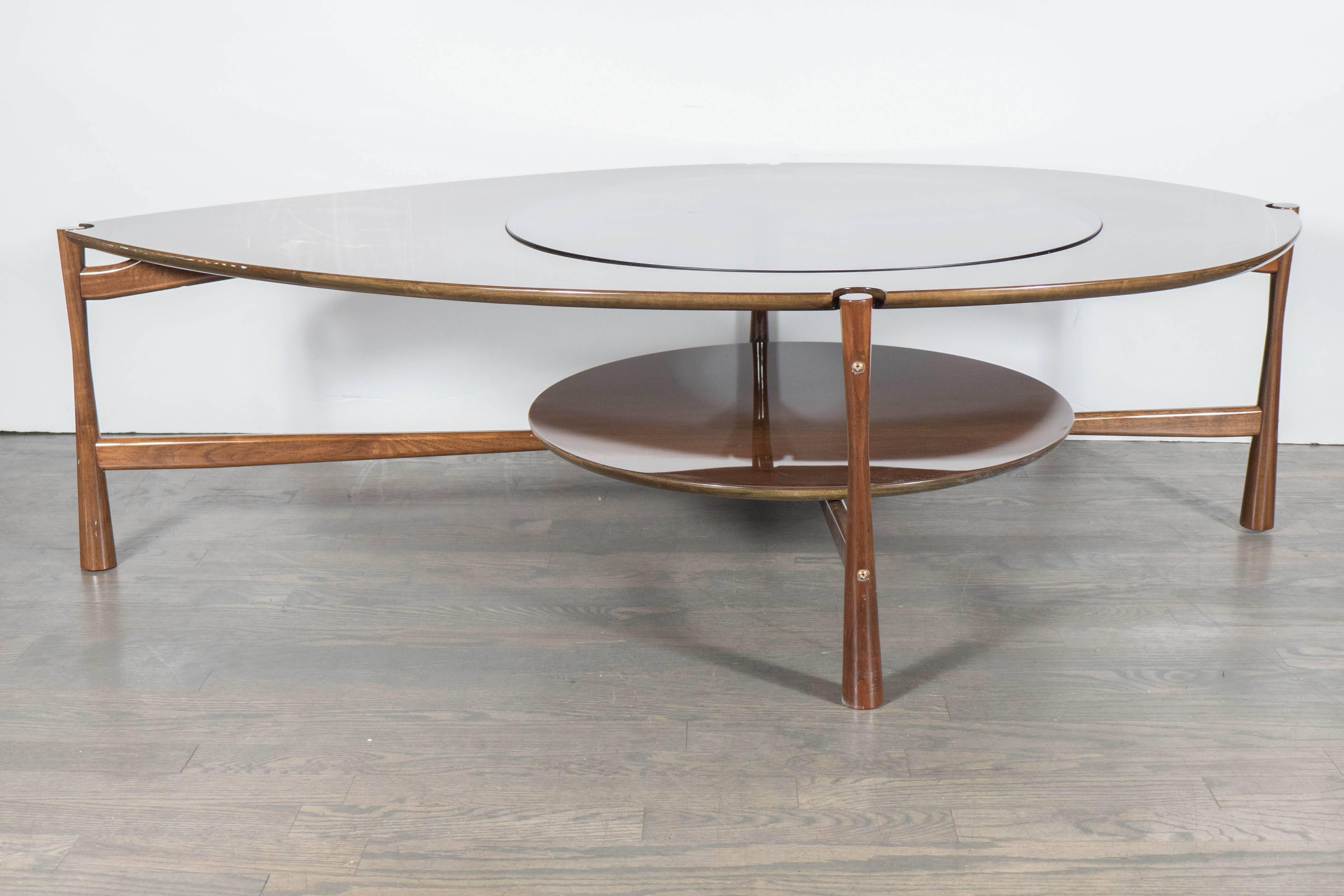 This sculptural Mid-Century Modernist cocktail table features a two-tier design in walnut, the top tier being a large tear drop shape with inset smoked glass center positioned directly over the circular lower tier, the two tiers are supported by