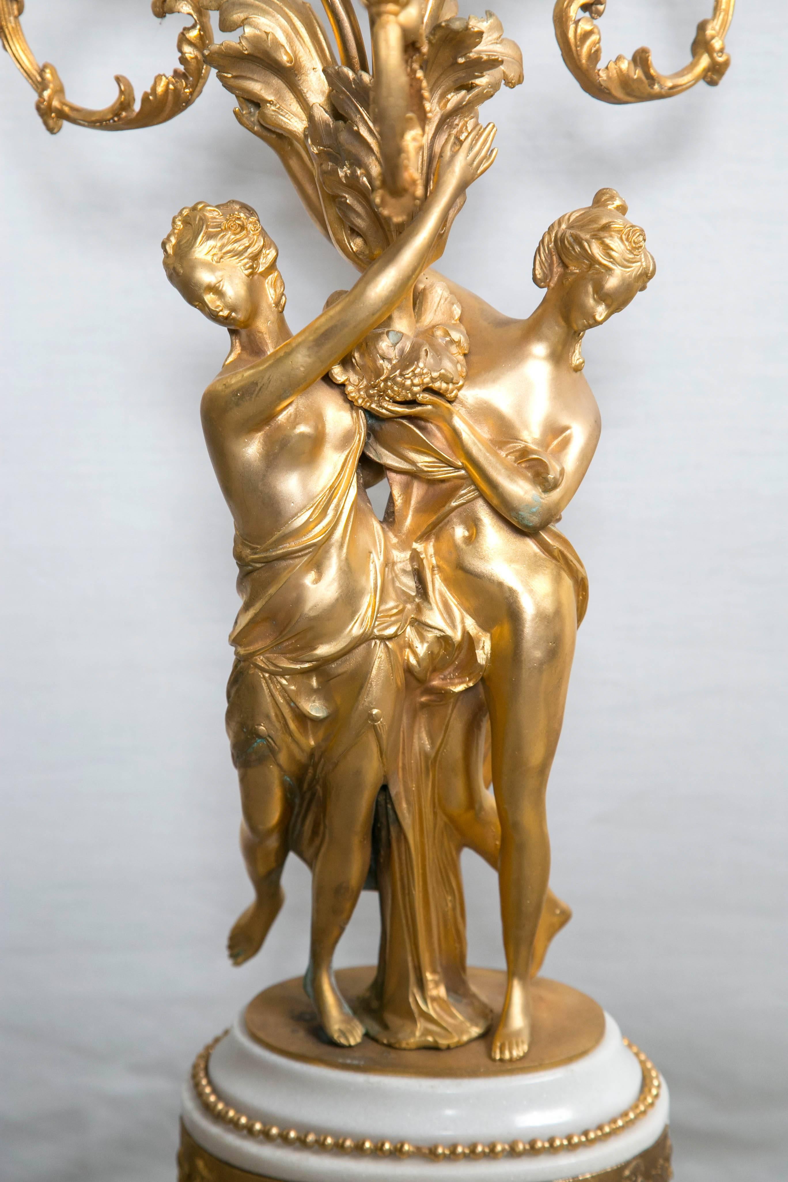 Each white marble base with gilt bronze mounts and feet. A band of frolicking putti, above a ring of leaves and appliques. Two partially draped females hold up the gathered scrolling branches of the three lights and center.