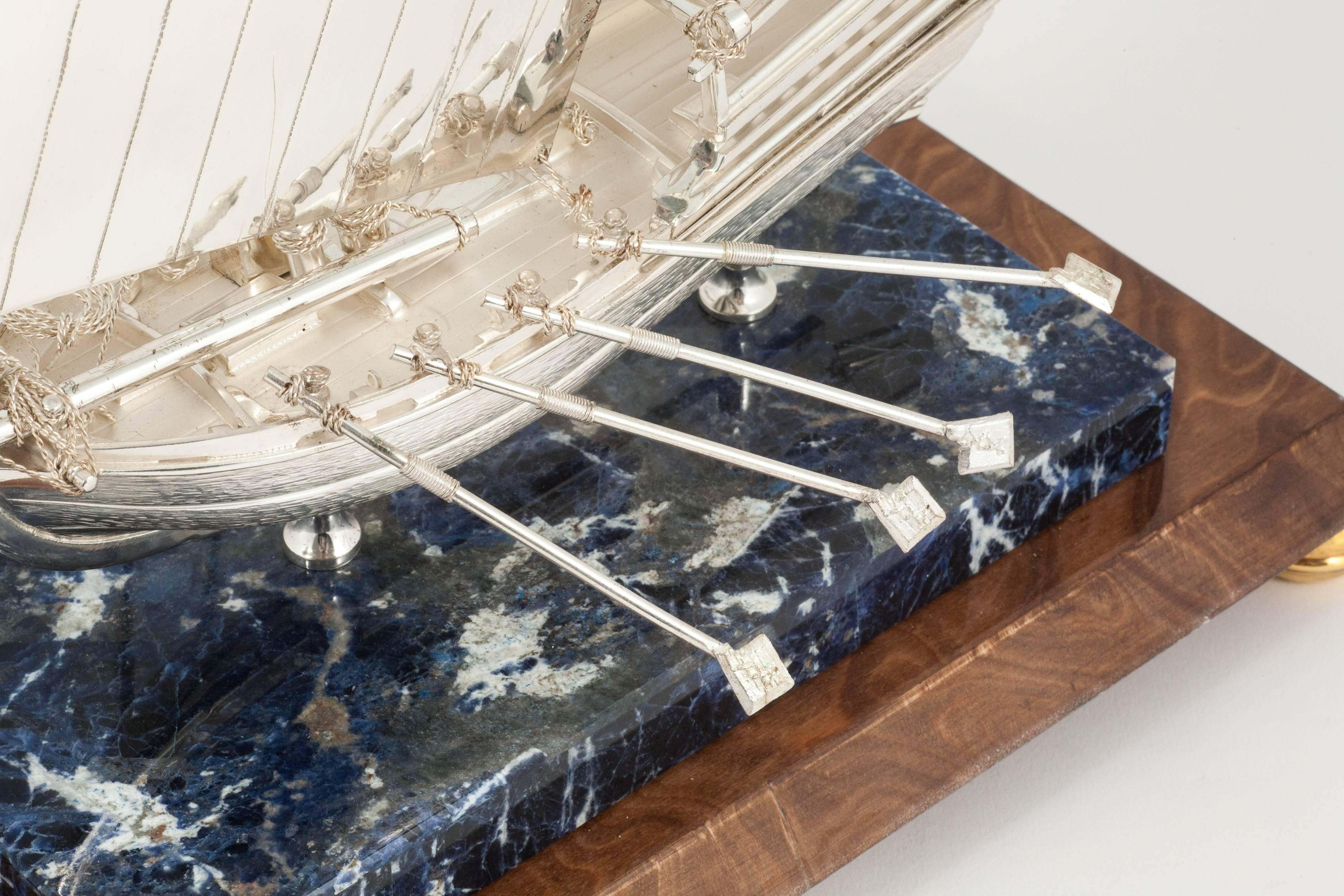 This scale silver model of a dhow is finely handmade with realistic rigging and deck detail. It is made of substantial heavy gauge silver with cast sections and four oars on each side. The stern of this yacht carries an enameled flag of the United