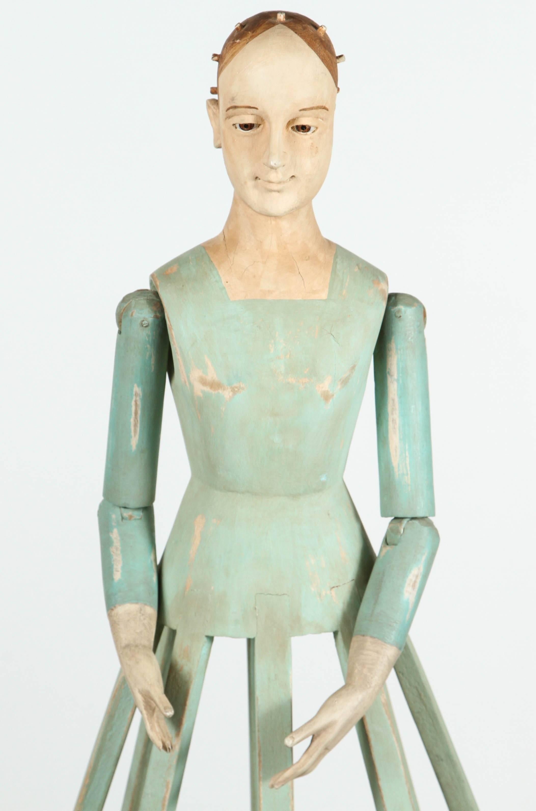 Vintage handcrafted Santo Mannequin.

Wooden turquoise dress.
Three angels on bottom base.
Wooden platform.

Mid-19th century bastidor (cage) santo with inset glass eyes, articulated arms jointed at the elbow and shoulder, together with the