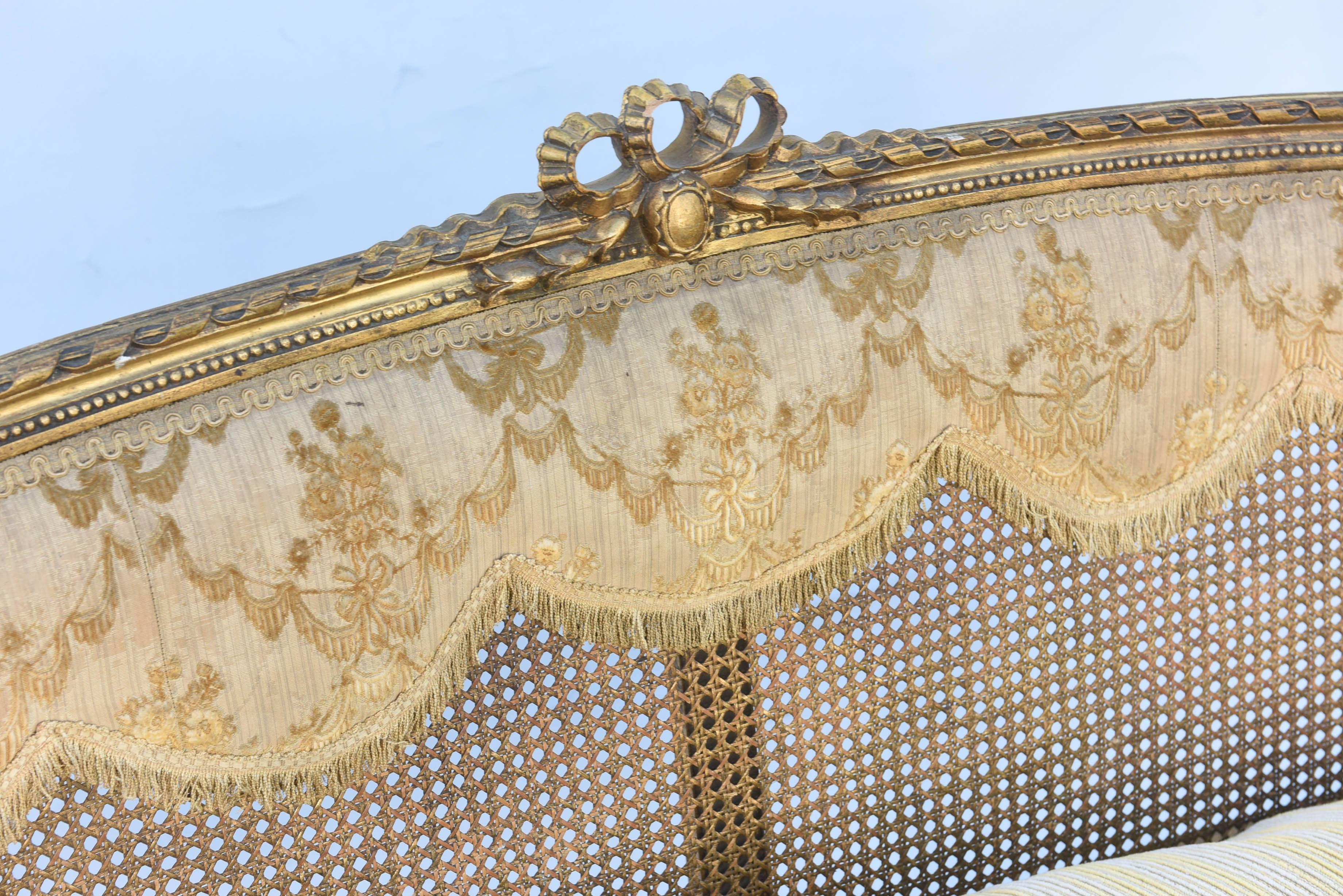 Louis XVI settee, of carved giltwood, having a bowed, rope-carved crestrail, centered by pierced ribbons, its caned back decorated by original embroidered silk drape, flanked by downswept, padded armrests, on spiral carved standards, its oval seat