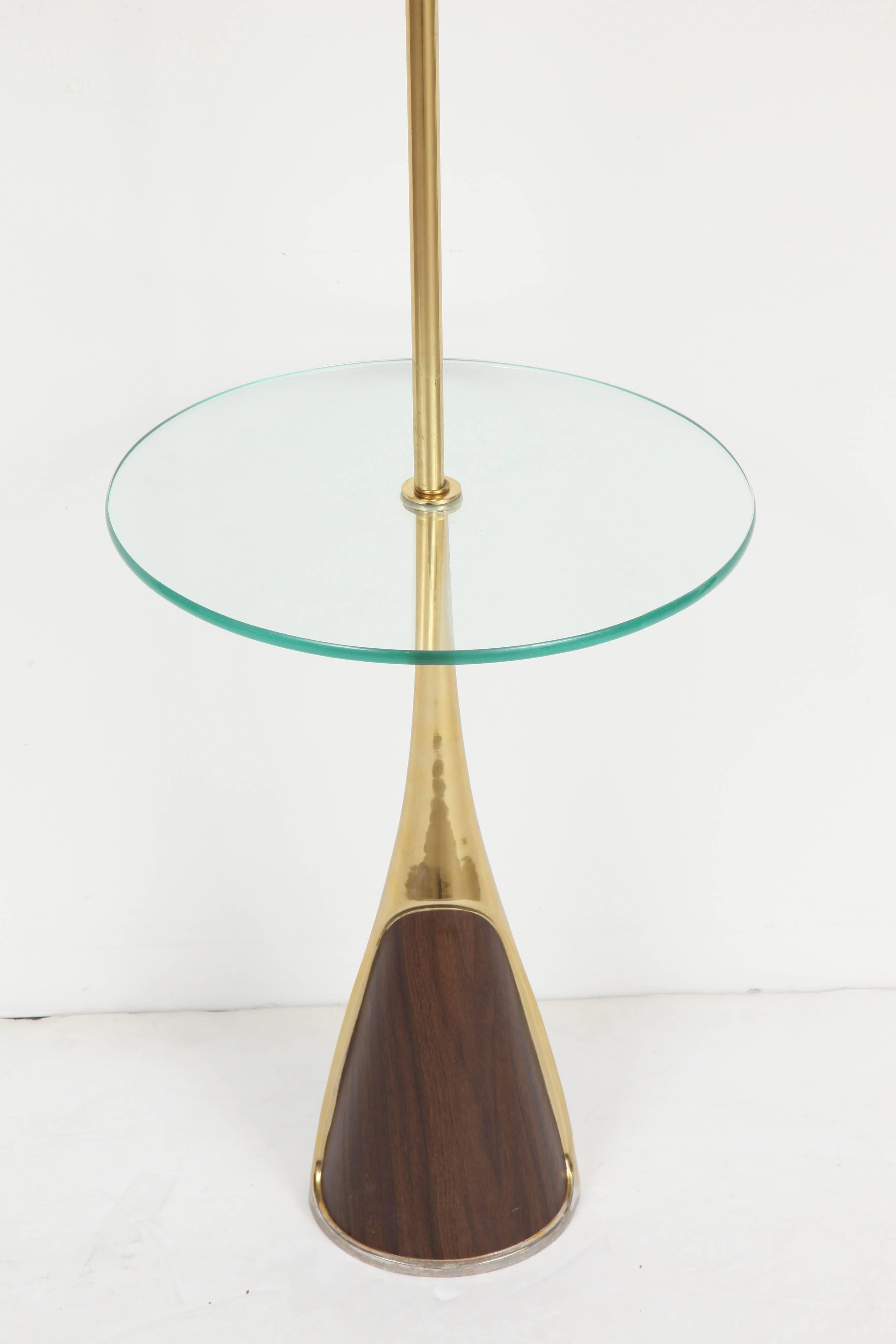 A floor lamp with round glass table shelf by Laurel. USA, circa 1950.  Features a polished brass and wood veneer base supporting a glass shelf; complemented by an off-white paper drum shade and original finial.  

Takes a standard bulb, 100 watts