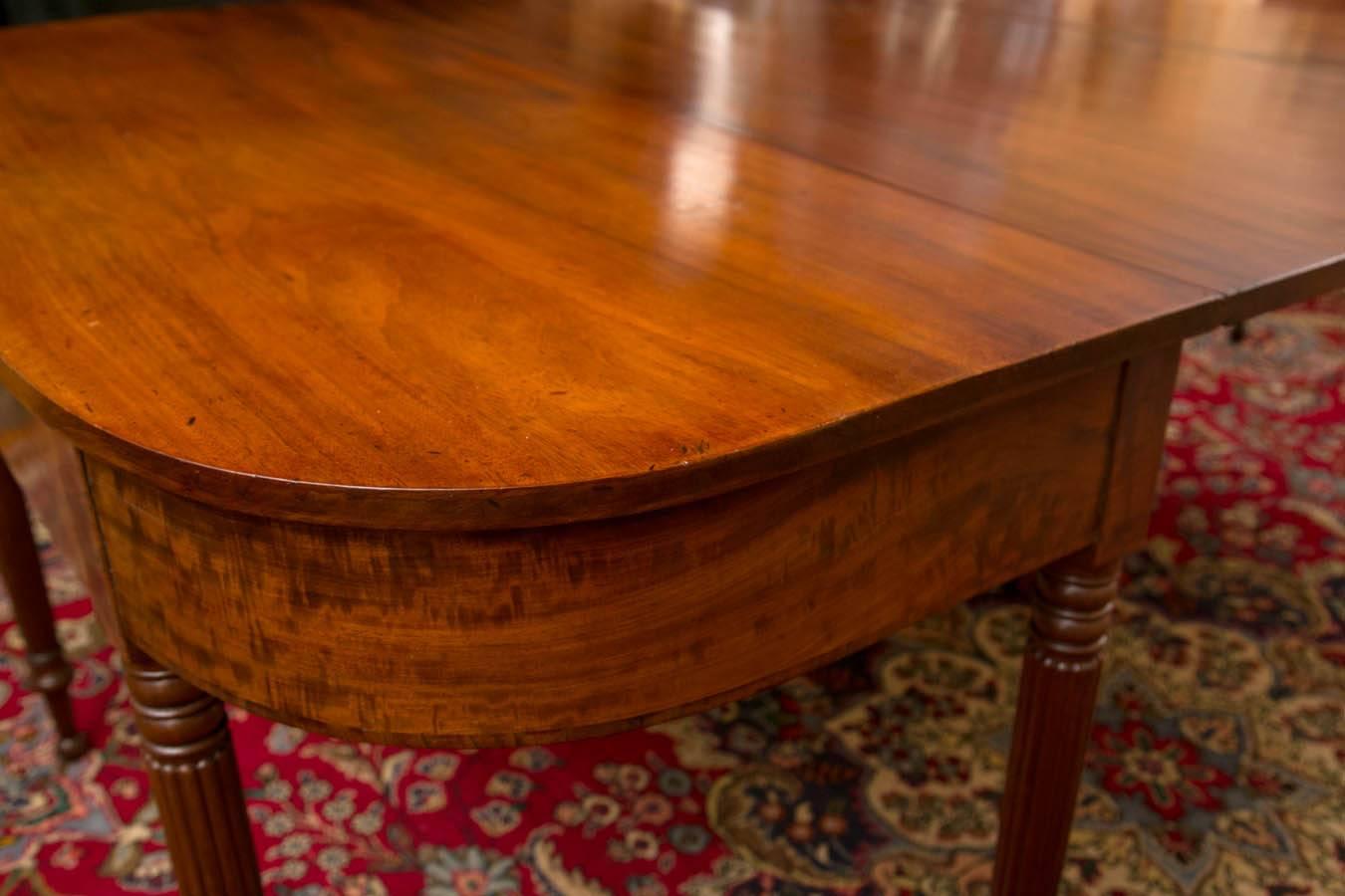 Unusual Sheraton mahogany two-part dining table, comprising two sections, each with D-Form top with blocked end centering the wide apron of figured mahogany. Each section with rectangular hinged leaf at the rear, raised on five reeded ring turned
