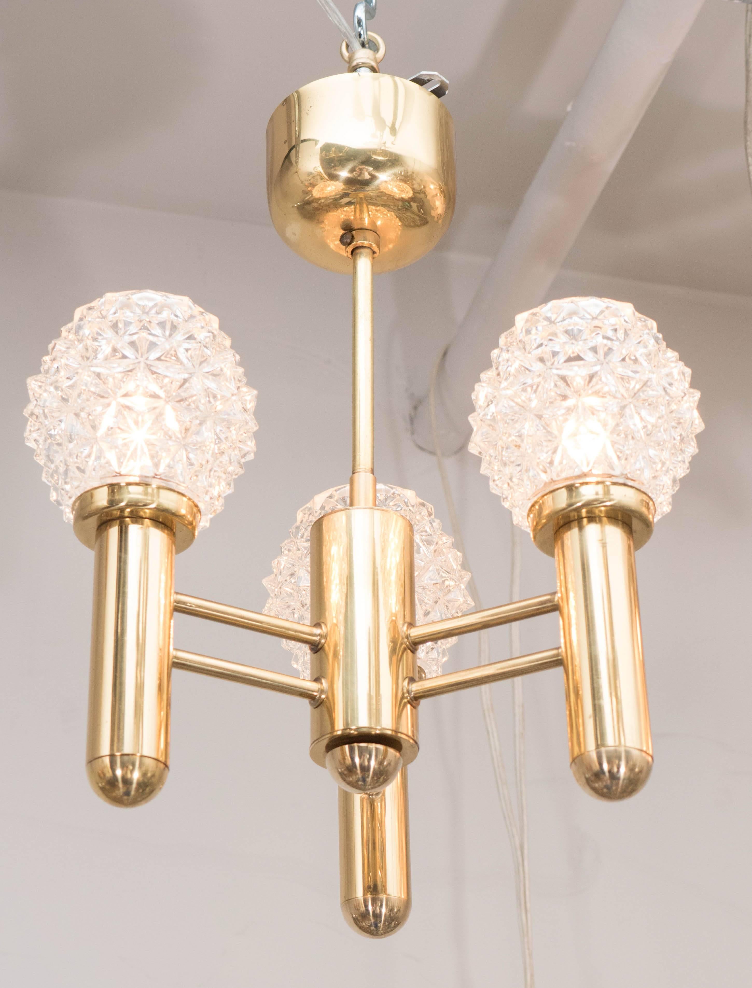 This elegant Mid-Century Modern chandelier was realized in Germany, circa 1960. It features a polished brass frame consisting of three cylindrical bodies adjoined to a central stem of the same form via horizontal cylindrical supports, all in