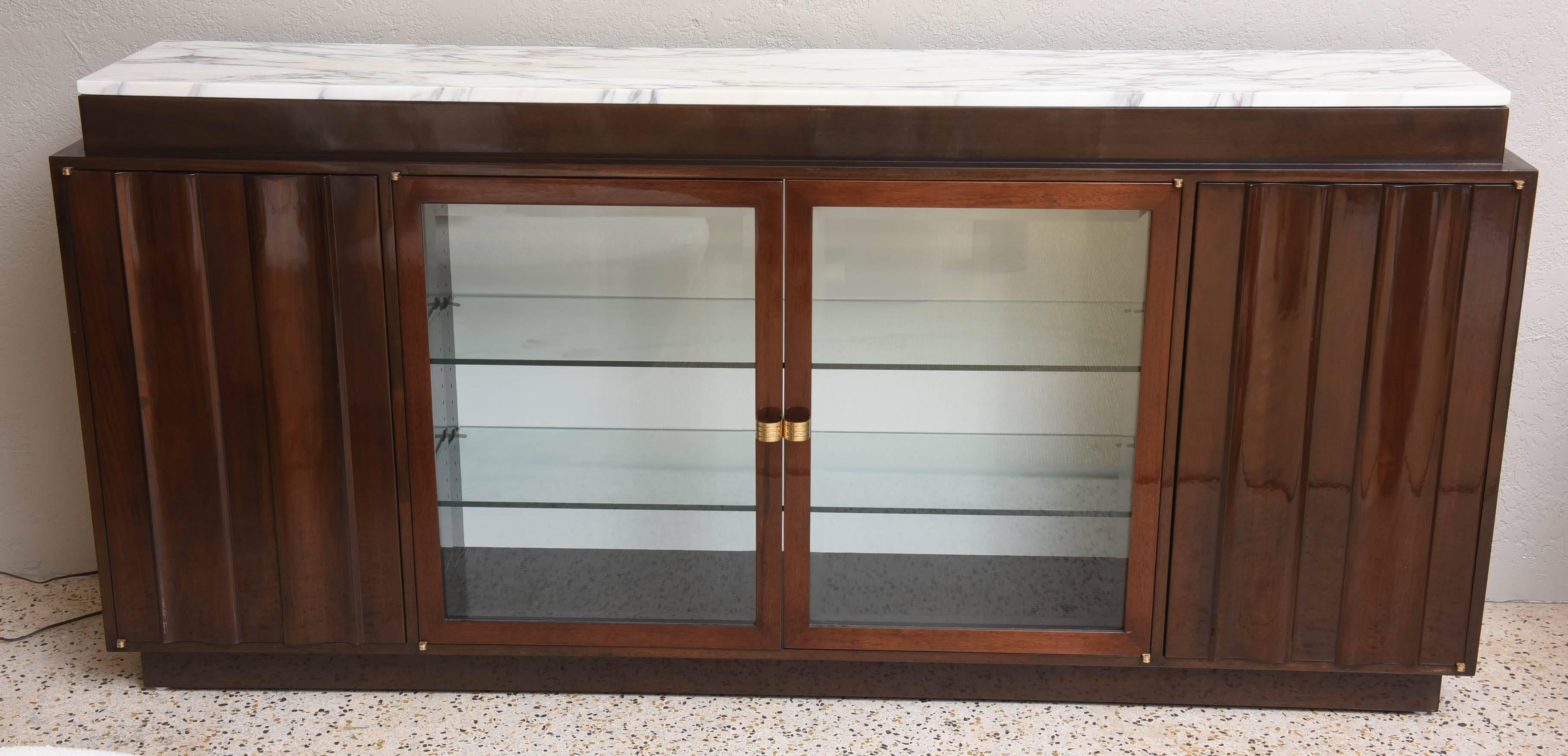 Tall fluted walnut buffet with marble top by Tommi Parzinger. Glazed double-doored lighted section with two glass shelves. Side cabinets with one shelf each and polished solid brass hardware. Part of a two-piece South Florida commission.