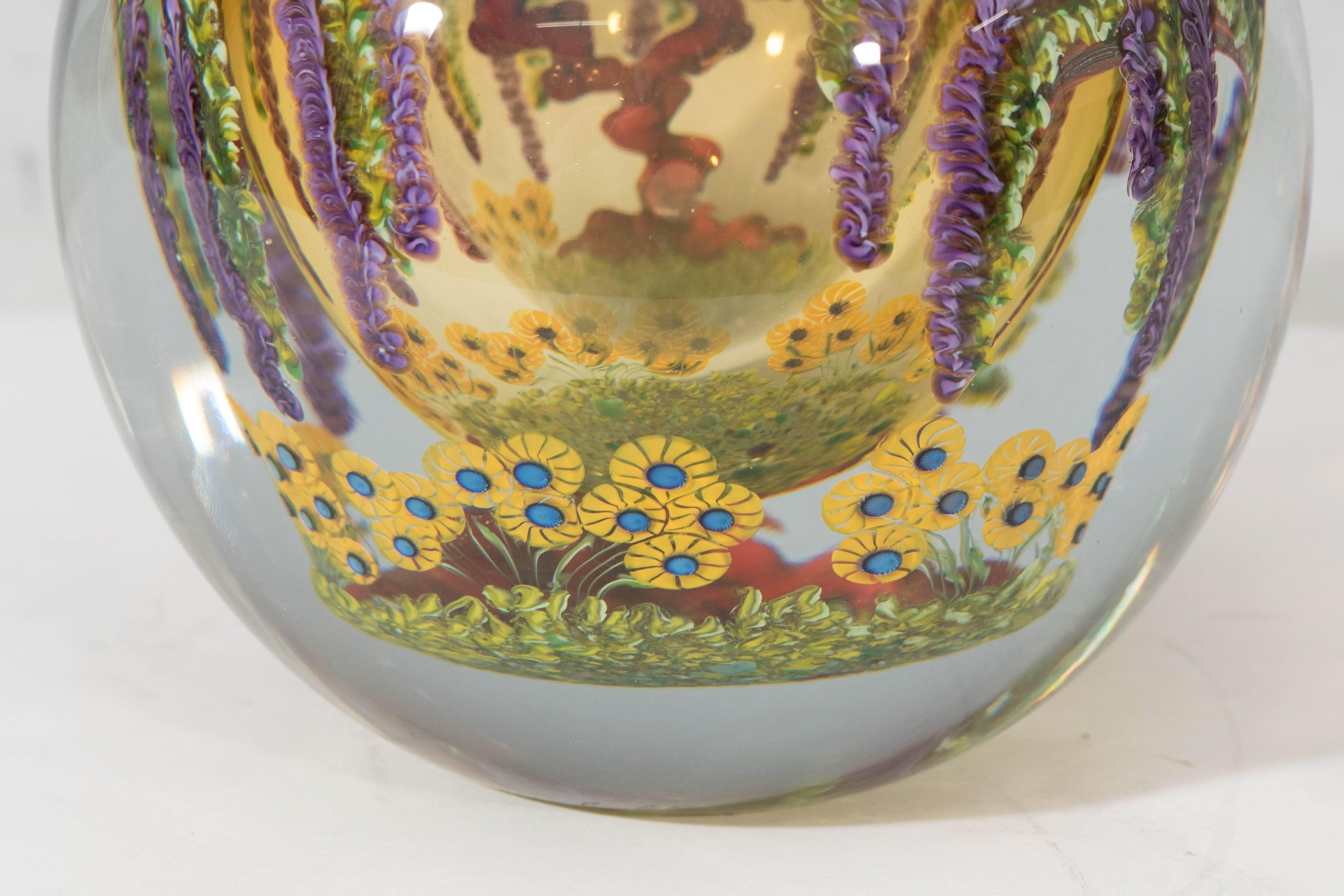 A handblown glass vase, created in 1997 by artist Chris Heilman, composed of intricate compound layers, clear over warm amber, detailed with hanging wisteria and twisting branches, above beds of bright yellow flowers. Markings include [Chris