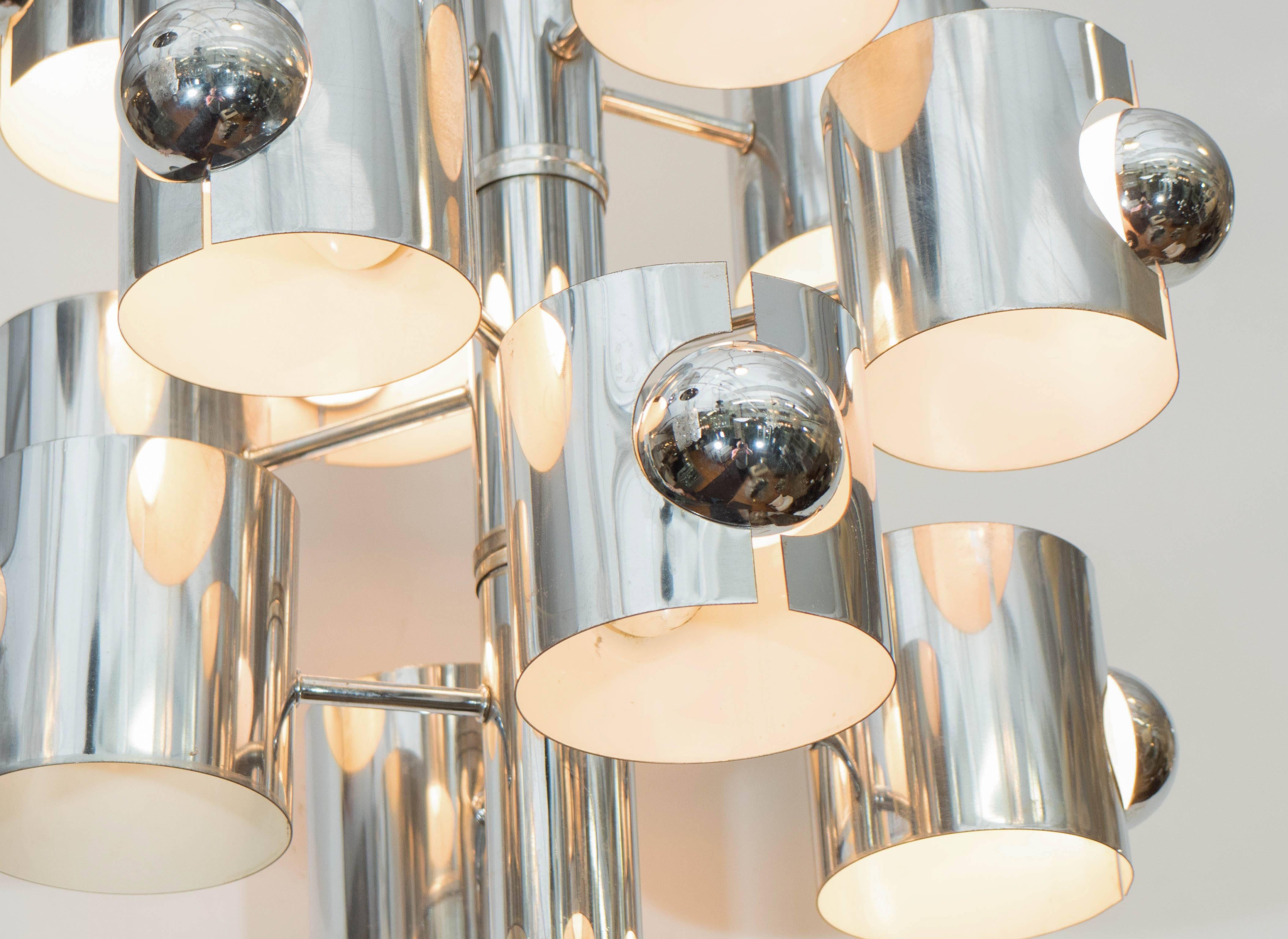 A vintage, highly modernistic chrome-plated chandelier by Gaetano Sciolari, produced circa 1960s-1970s, which includes 12 lights, dispersed into three tiers, with cylindrical cut-out shades, each exposing the bulb, the interiors enameled white.