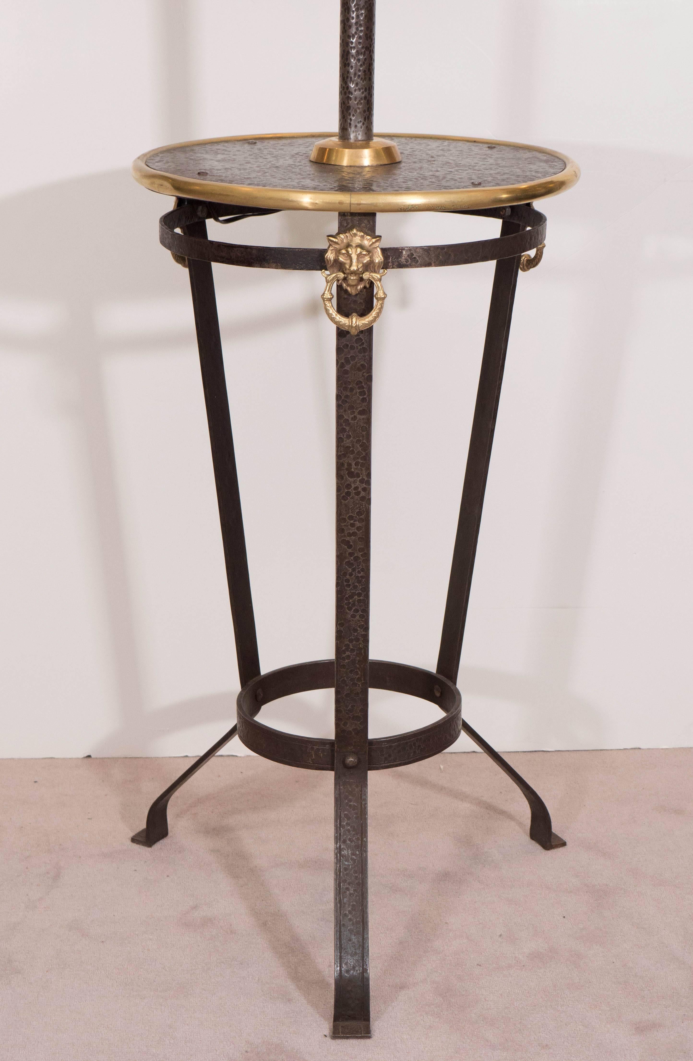 This incredibly unique Arts & Crafts table lamp, produced circa 1910, comes in hammered iron on tripod legs, with a round table, trimmed along the edge in bright brass, decorated with three lion heads with rings; the table measures 17