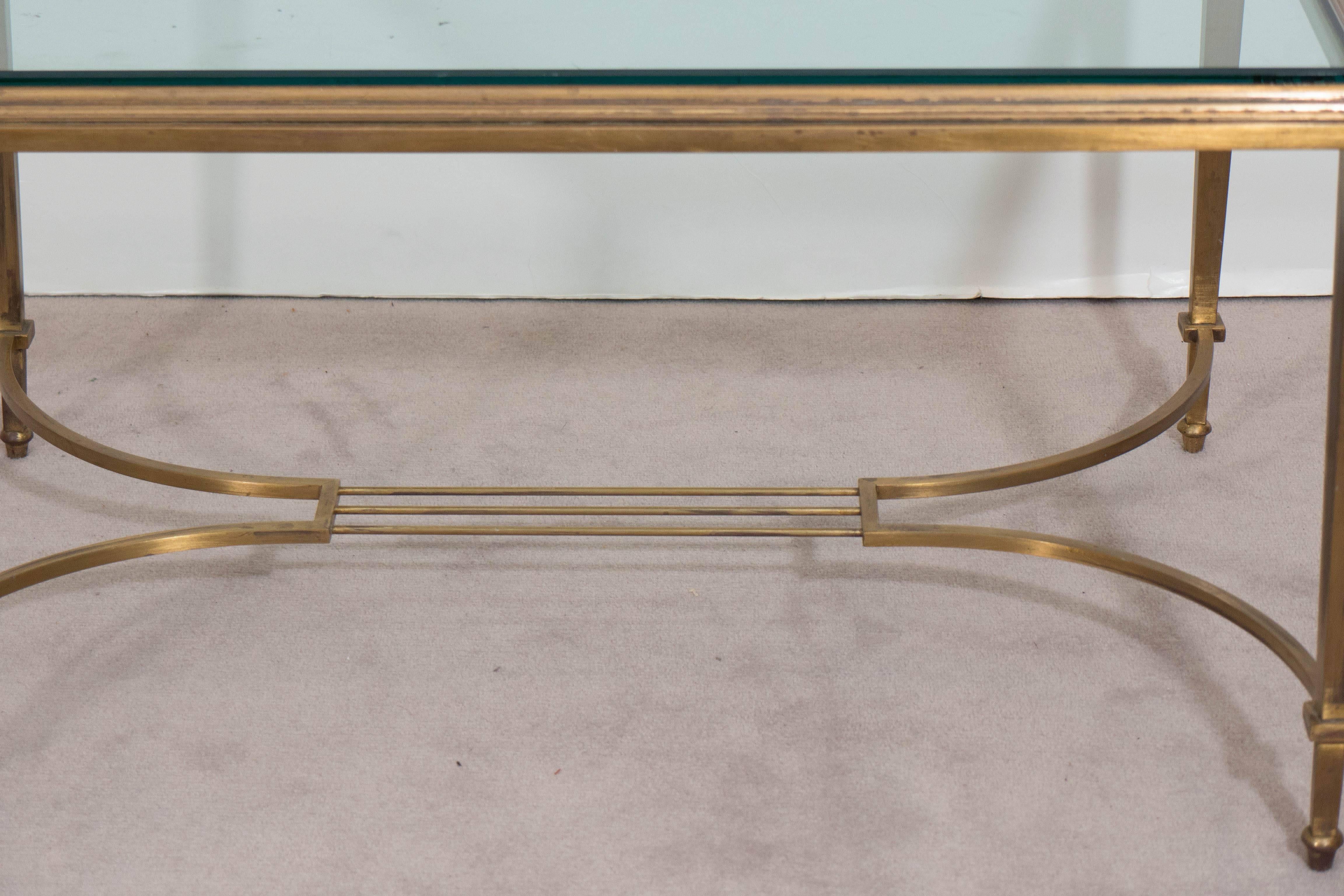 A vintage coffee table designed in the neoclassical taste circa 1950s, with glass top on a beveled brass frame and tapered legs, with harp form stretcher. This piece is in good condition, with age appropriate wear to finish, as well as minor