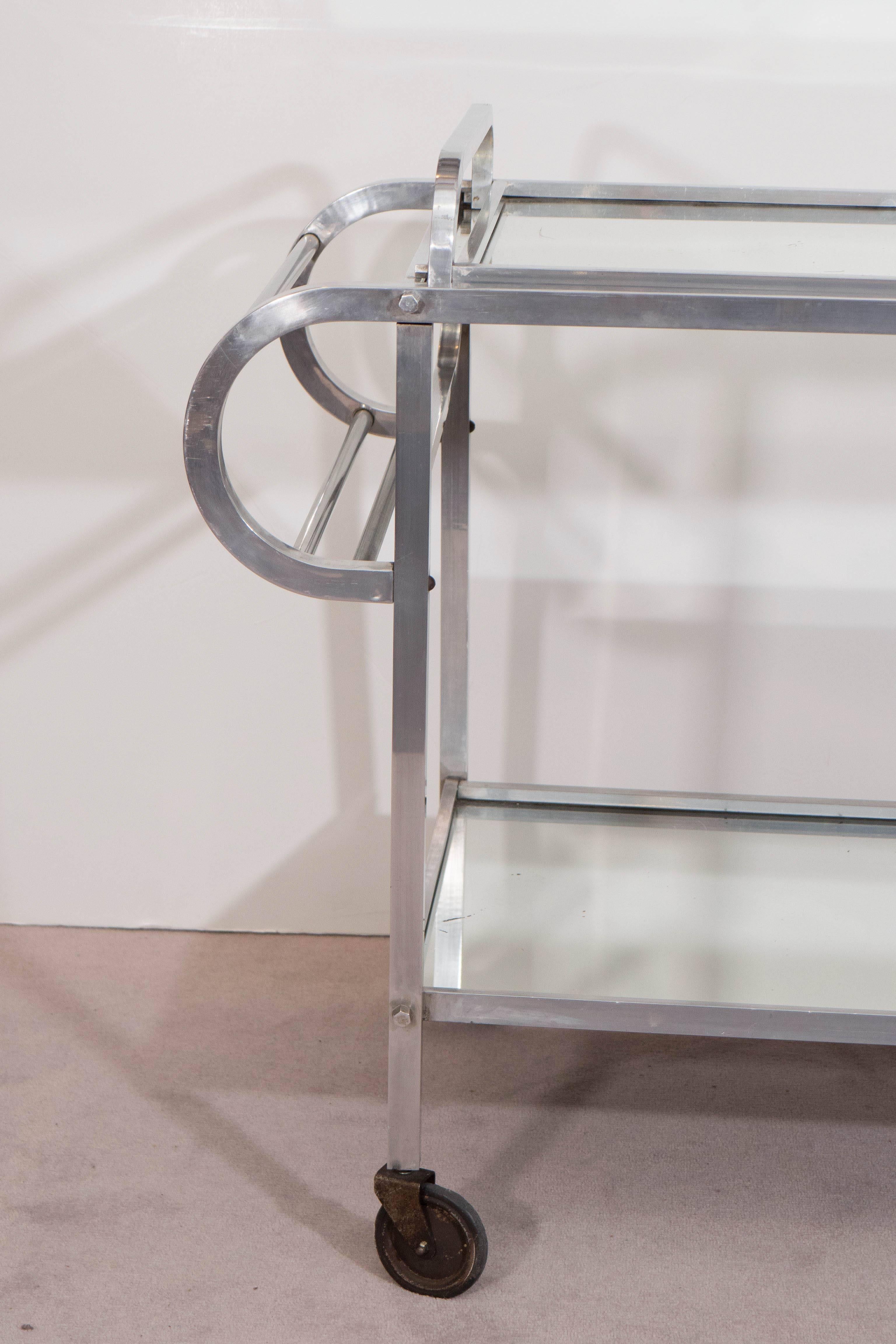 This beautifully linear bar cart, produced in France circa 1930s, comes in a brushed aluminum frame, flanked by two rounded push handles, with two shelves with inset mirrors, on wheels; included is a matching serving tray. Despite some loss of