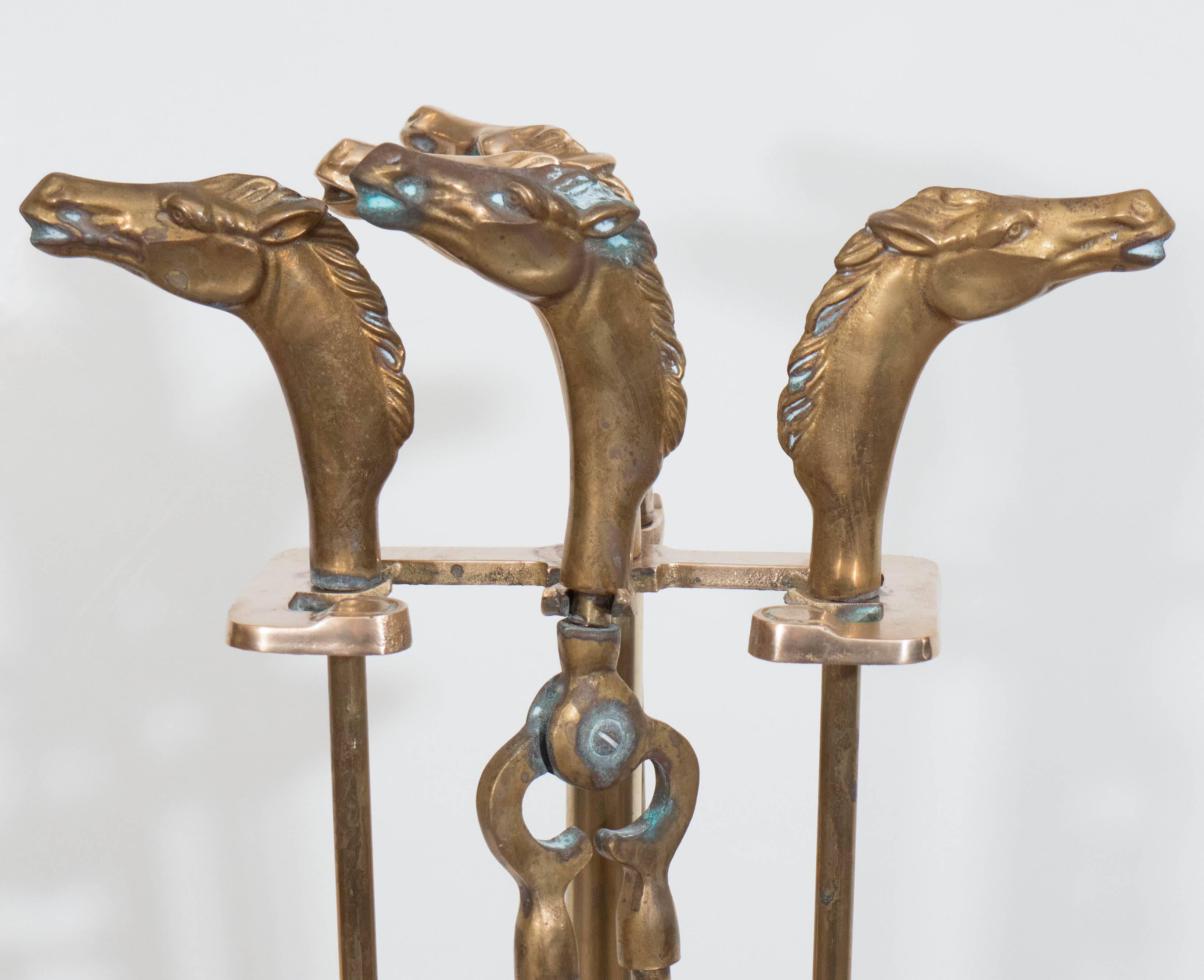 This set of brass fireplace tools, produced circa 1950s, includes broom, tongs, shovel and poker on elaborate stand with gallery to the base, each with beautifully detailed horse head motif as handle. Despite some loss of broom bristles, as well as