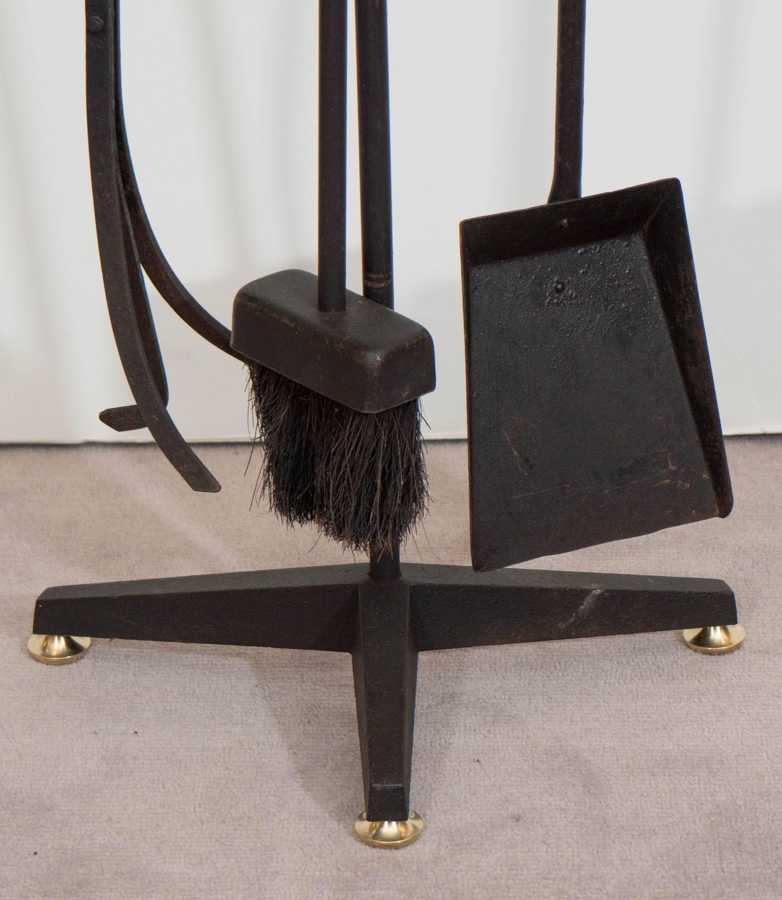 This set of fireplace tools in wrought iron, designed by Donald Deskey circa 1950s for Bennett Co., includes shovel, broom and a pair of tongs, handles detailed with brass ball finial, on a tripod stand. Despite some rusting, as well as presence of