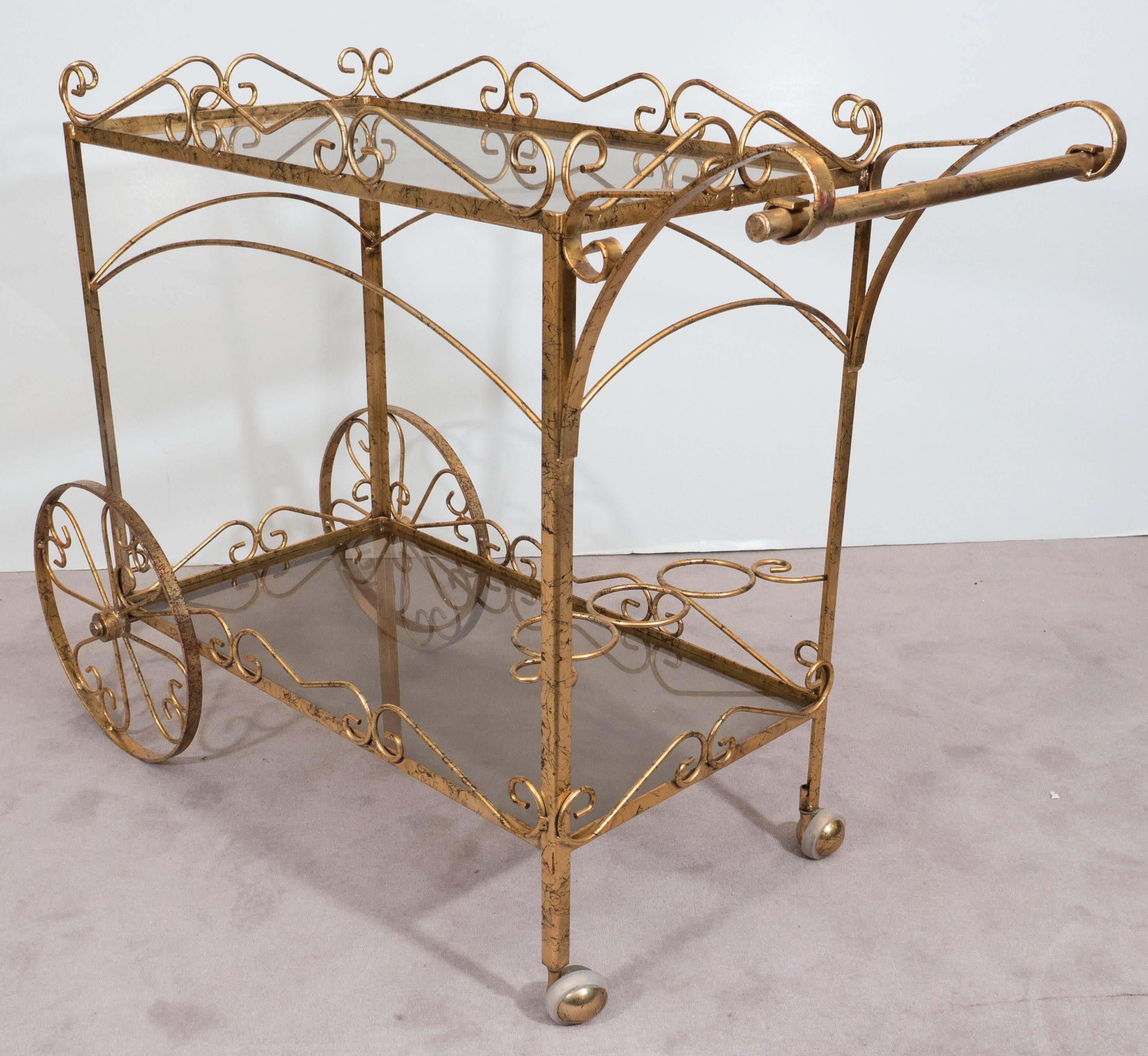 A vintage bar cart, with beautiful scrollwork frame in gilded metal, including two shelves of smoked glass and three bottle holders, on caster and spoke wheels. Very good vintage condition, with age appropriate wear to the gilt finish.