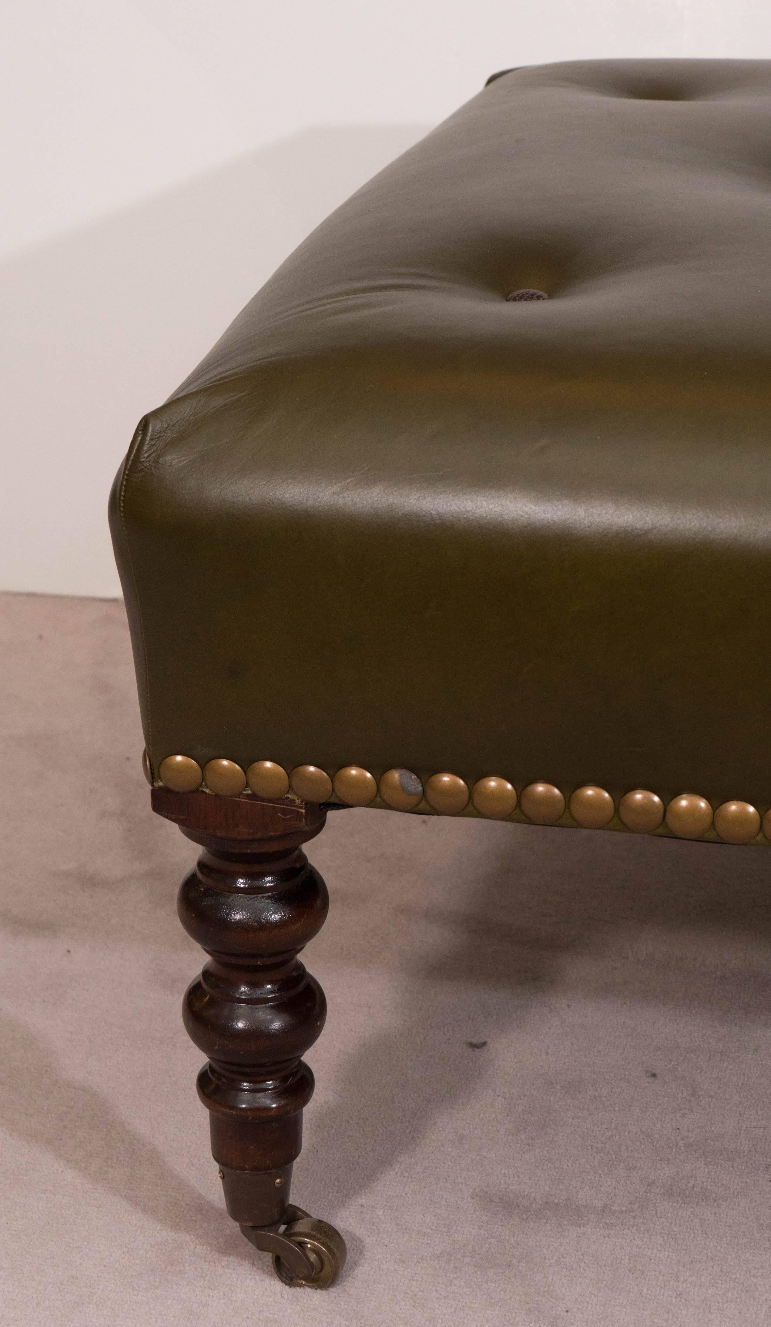 A vintage signature ottoman produced by George Smith, circa 1960s, with cushioned and button tufted seat in olive green leather, trimmed with brass nailheads, standing on bobbin turned legs in mahogany on caster wheels. This piece is in overall very